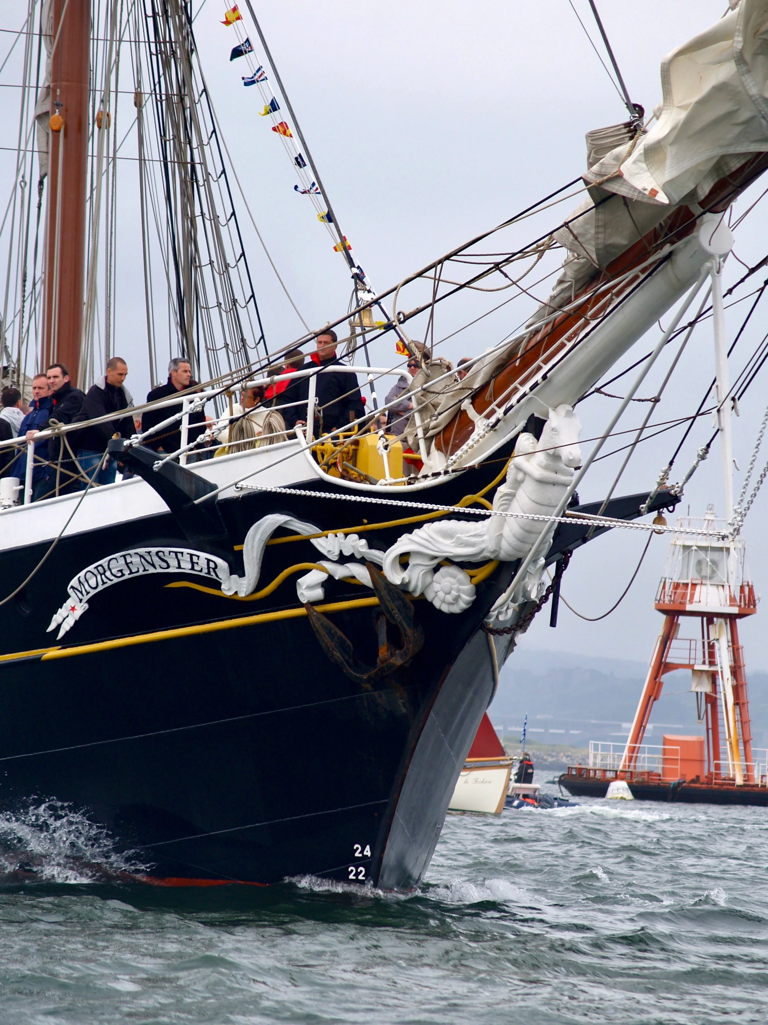 Un voilier en mer avec plusieurs personnes à bord, flotteurs et cordages visibles, fête nautique avec drapeaux, en arrière-plan une tour de signalisation maritime orange.