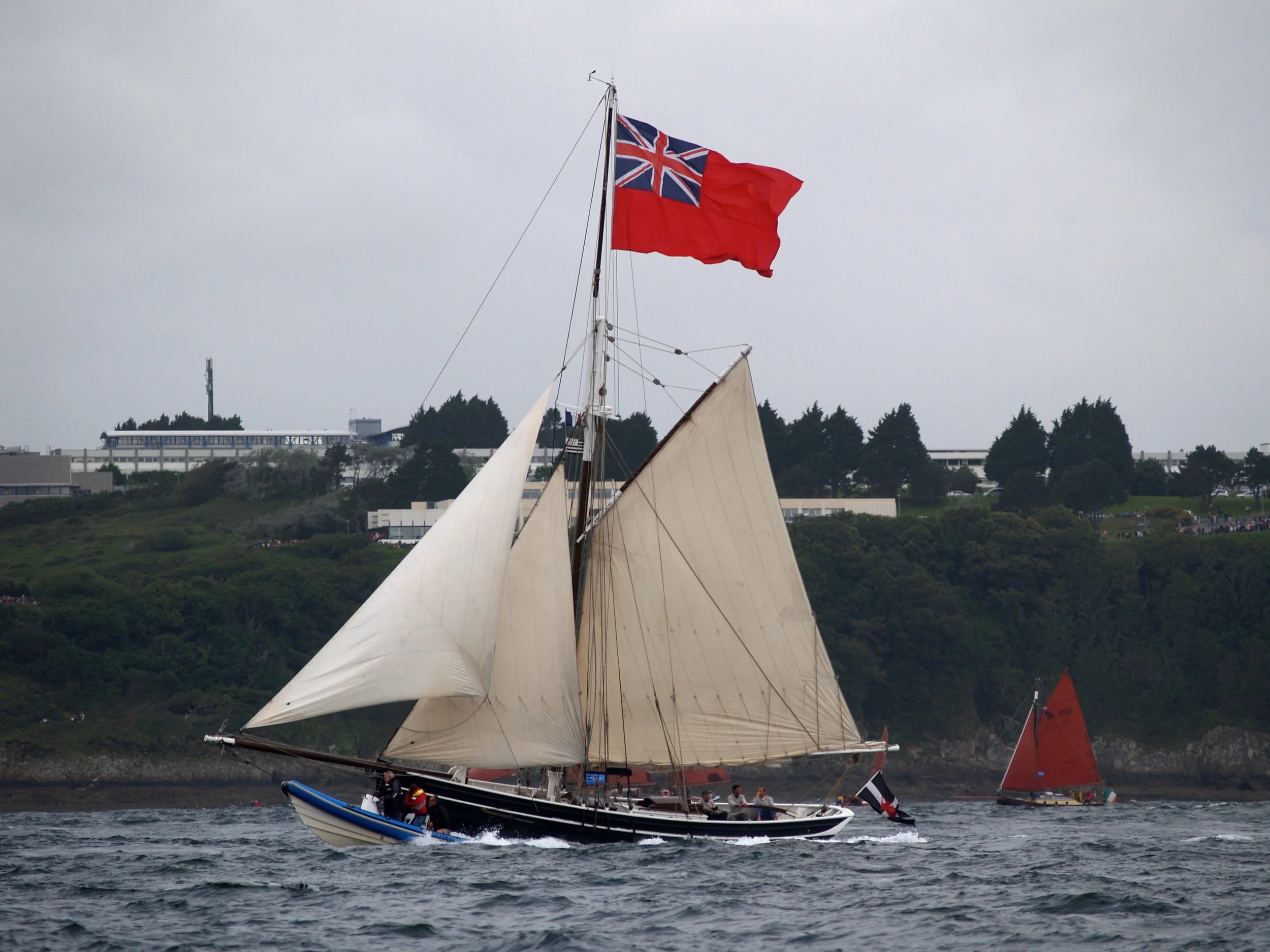 Un voilier avec ses voiles déployées navigue sur l'eau, avec un drapeau britannique et un drapeau rouge flottant en haut de la mâture, sous un ciel gris, en arrière-plan des bâtiments et des arbres.