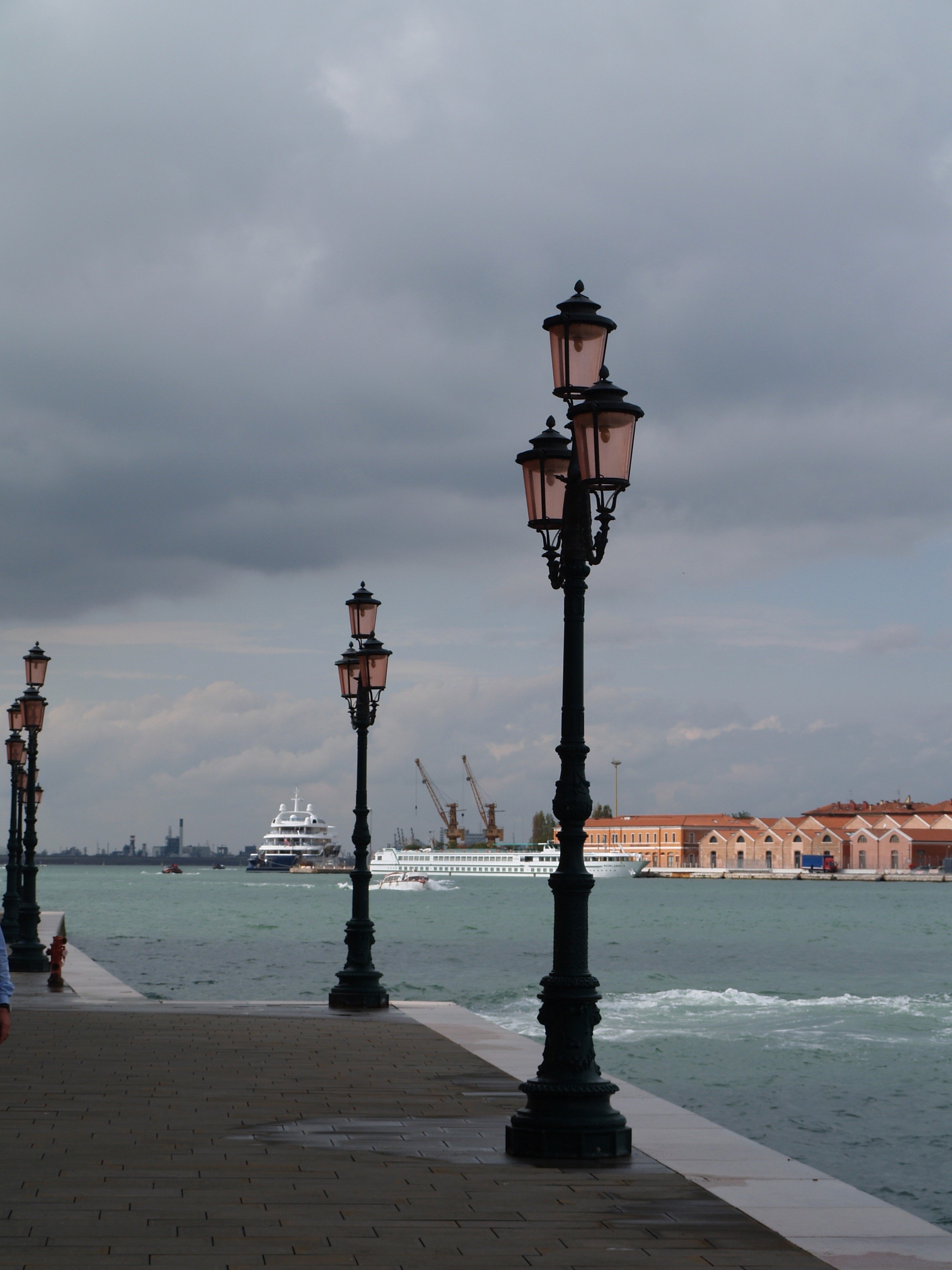 Un quai le long d'une rivière ou d'un port avec des lampadaires en fer noir, plusieurs bateaux de croisière ou yachts en mer, et un bâtiment industriel ou portuaire avec des grues de chantier en arrière-plan, sous un ciel nuageux.