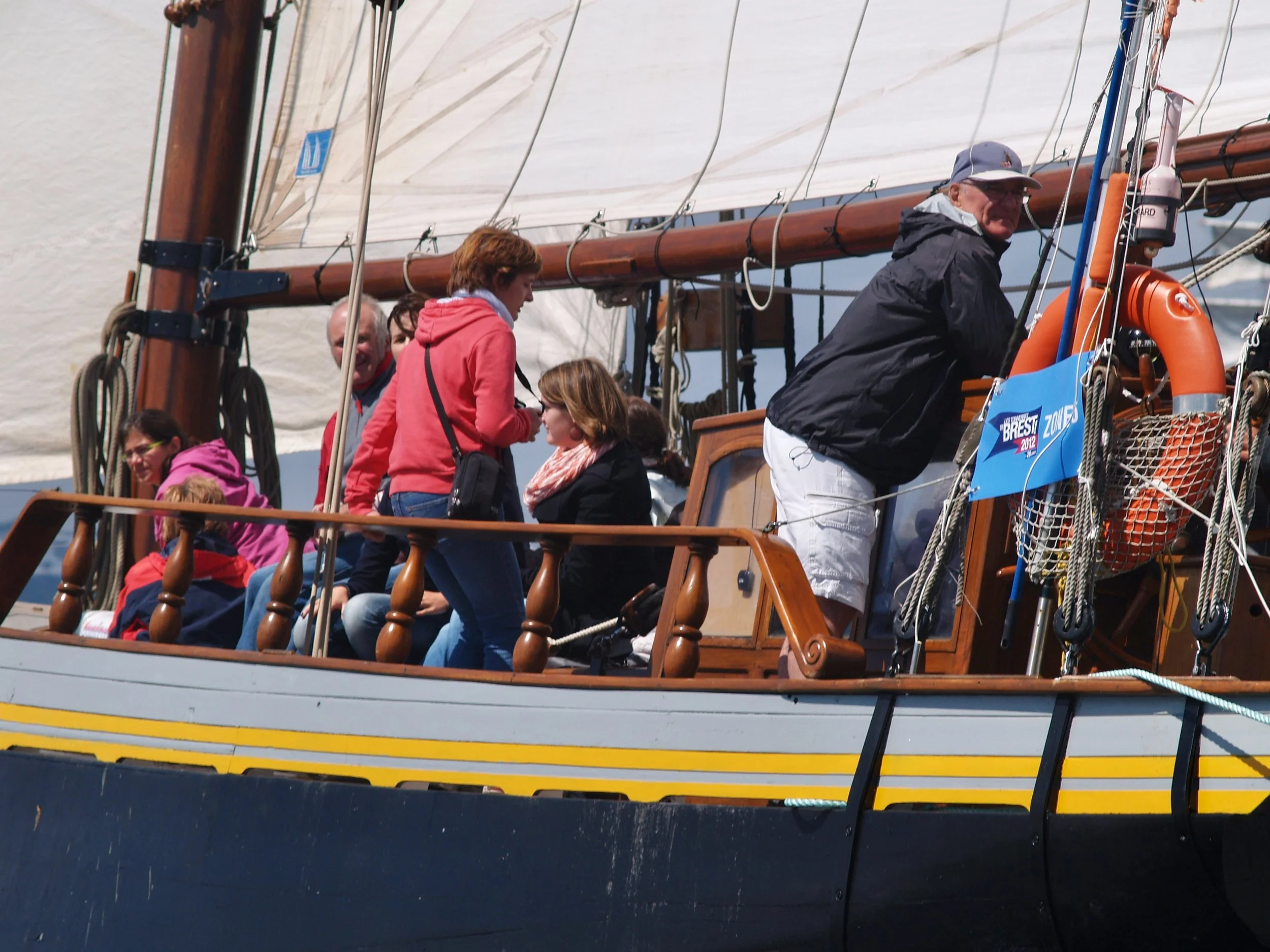 Groupe de personnes sur un voilier en mer, certains assis, d'autres debout, avec un marin à la barre. Le voilier a des voiles blanches et une coque bleue, avec un panneau indiquant "Brest 2012".
