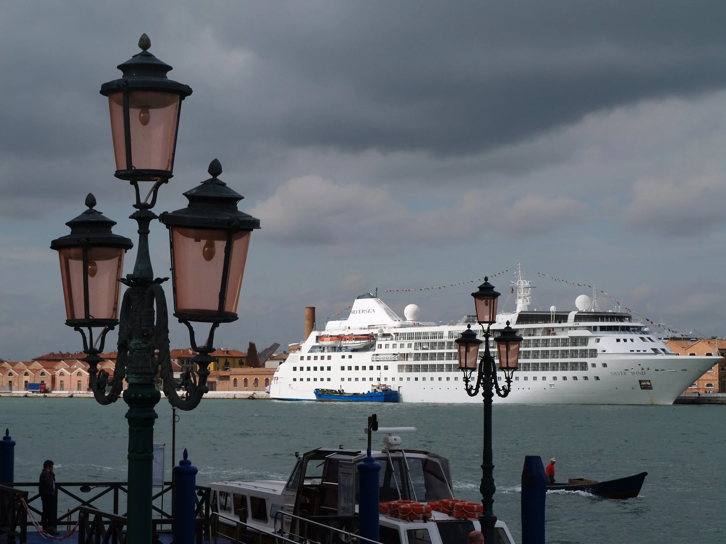 Un phare en métal noir avec des lanternes en verre teinté rose sur un quai au bord de l’eau, avec un grand paquebot nommé 'Silver Wind' dans le port, des bâtiments en arrière-plan et un ciel orageux.