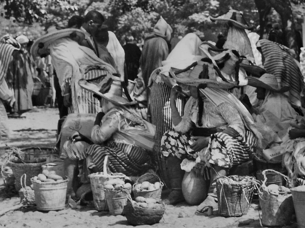 Marché en plein air avec des vendeurs de fruits et légumes, plusieurs personnes portant des chapeaux et des paniers.