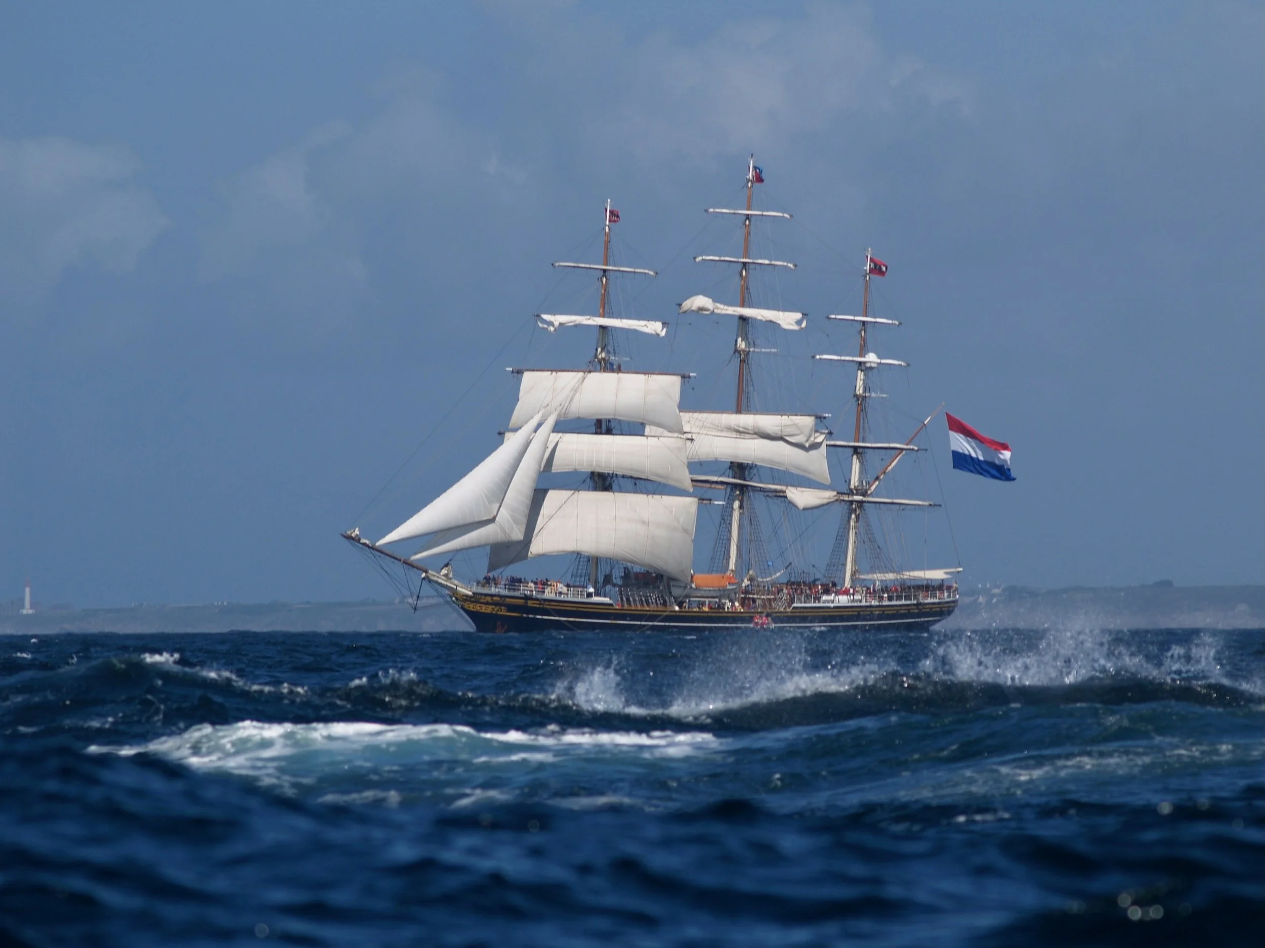 Un grand voilier avec des voiles blanches navigue sur la mer sous un ciel partiellement nuageux.