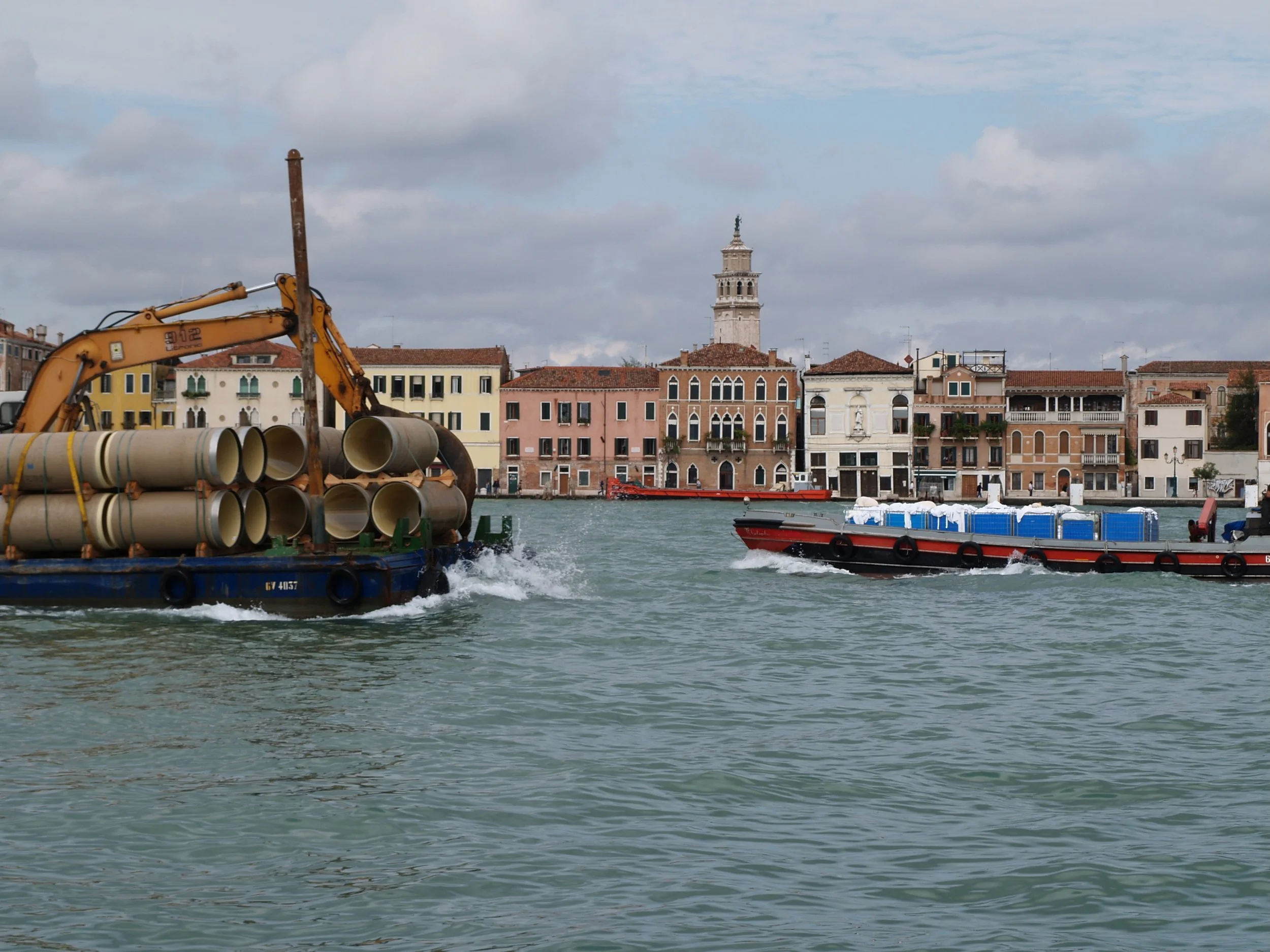 Vue sur un canal avec un bateau transportant des tuyaux en plastique, un autre bateau passe à côté. En arrière-plan, des bâtiments colorés et une tour avec une statue au sommet sous un ciel voilé.