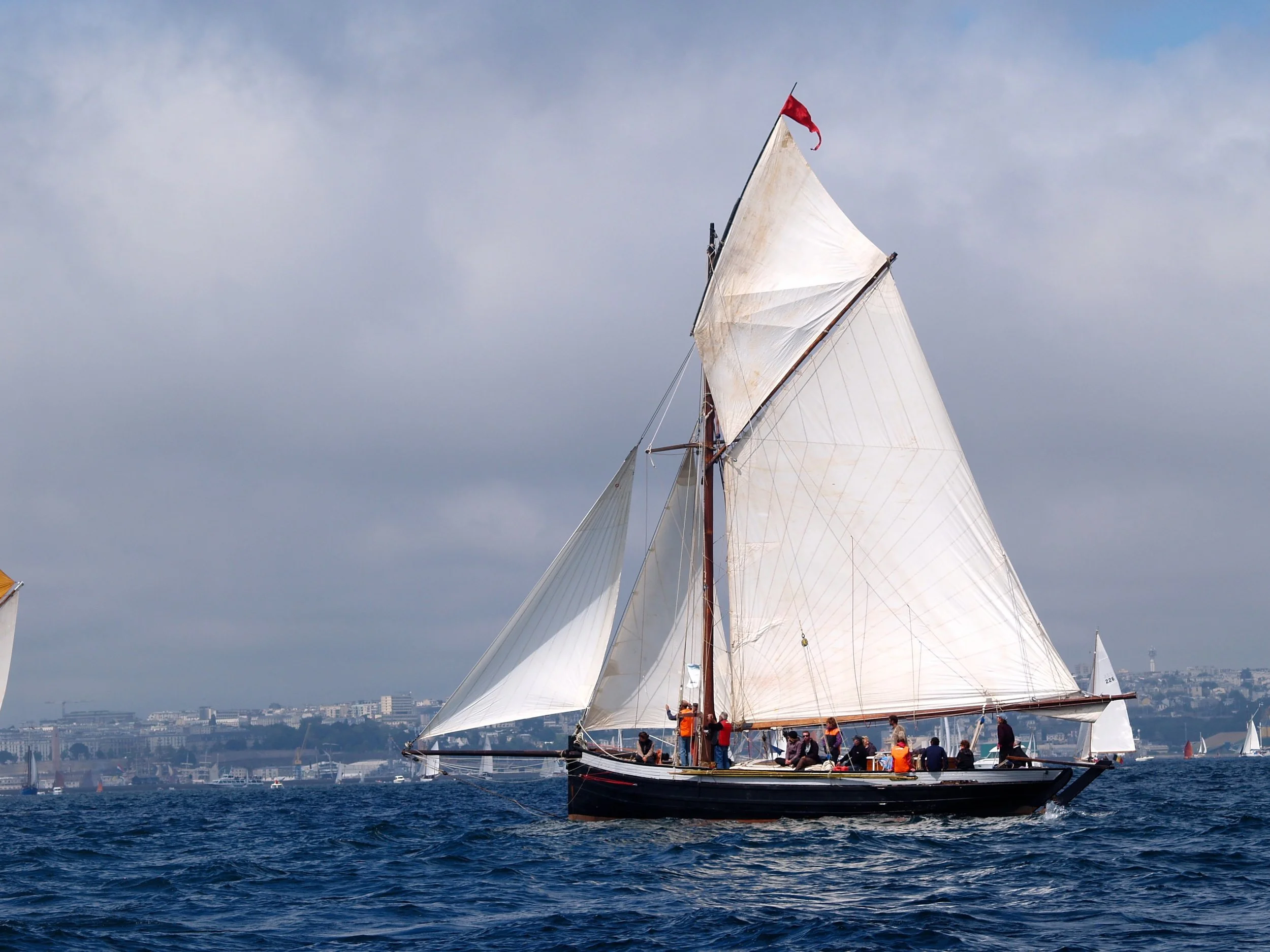 Un voilier avec des voiles blanches navigue sur l'eau, avec des hommes à bord et une ville en arrière-plan sous un ciel partiellement nuageux.