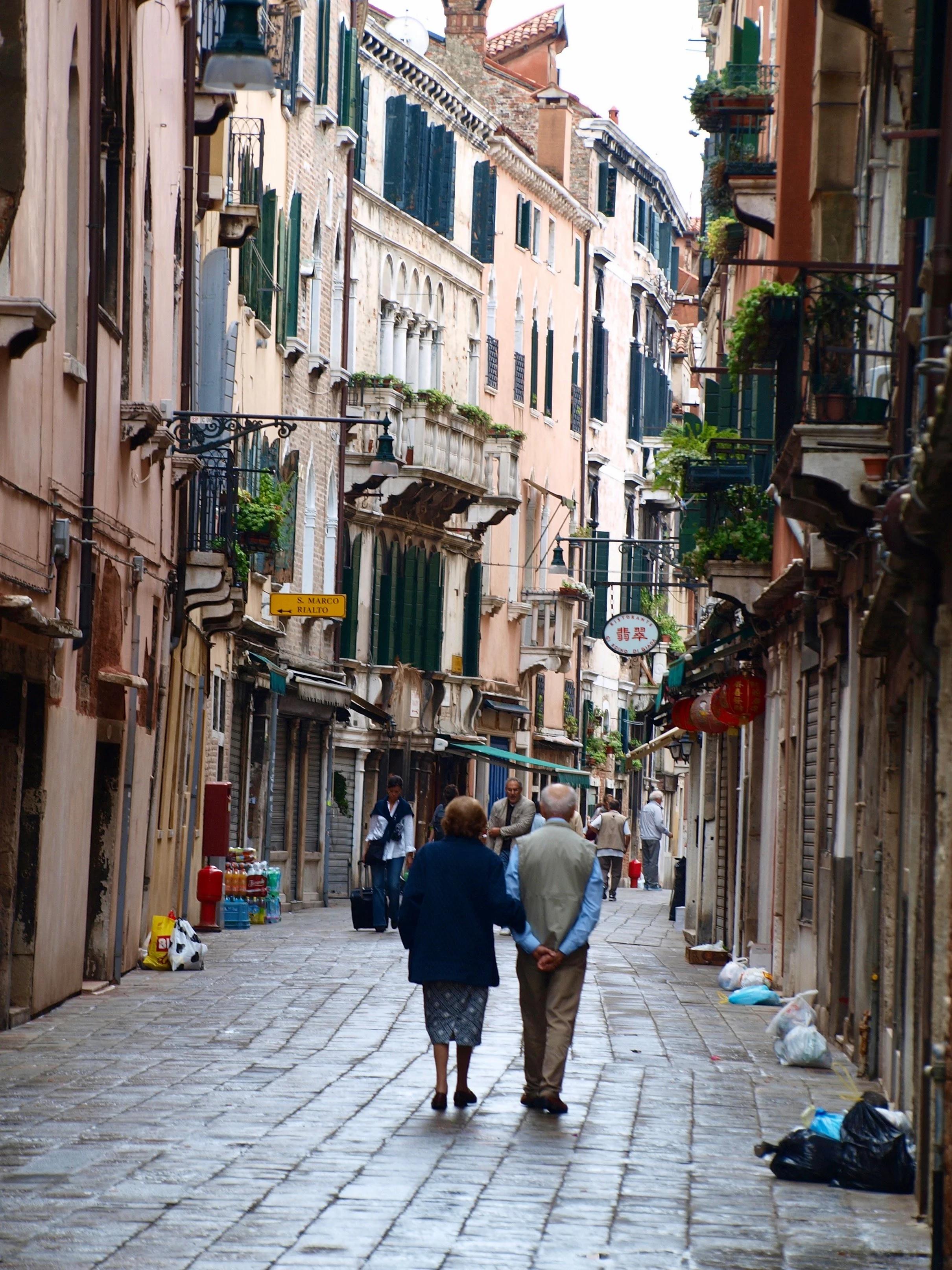 Une rue piétonne en ville avec des bâtiments colorés, des balcons avec des plantes et des lampes suspendues, et des personnes se promenant.