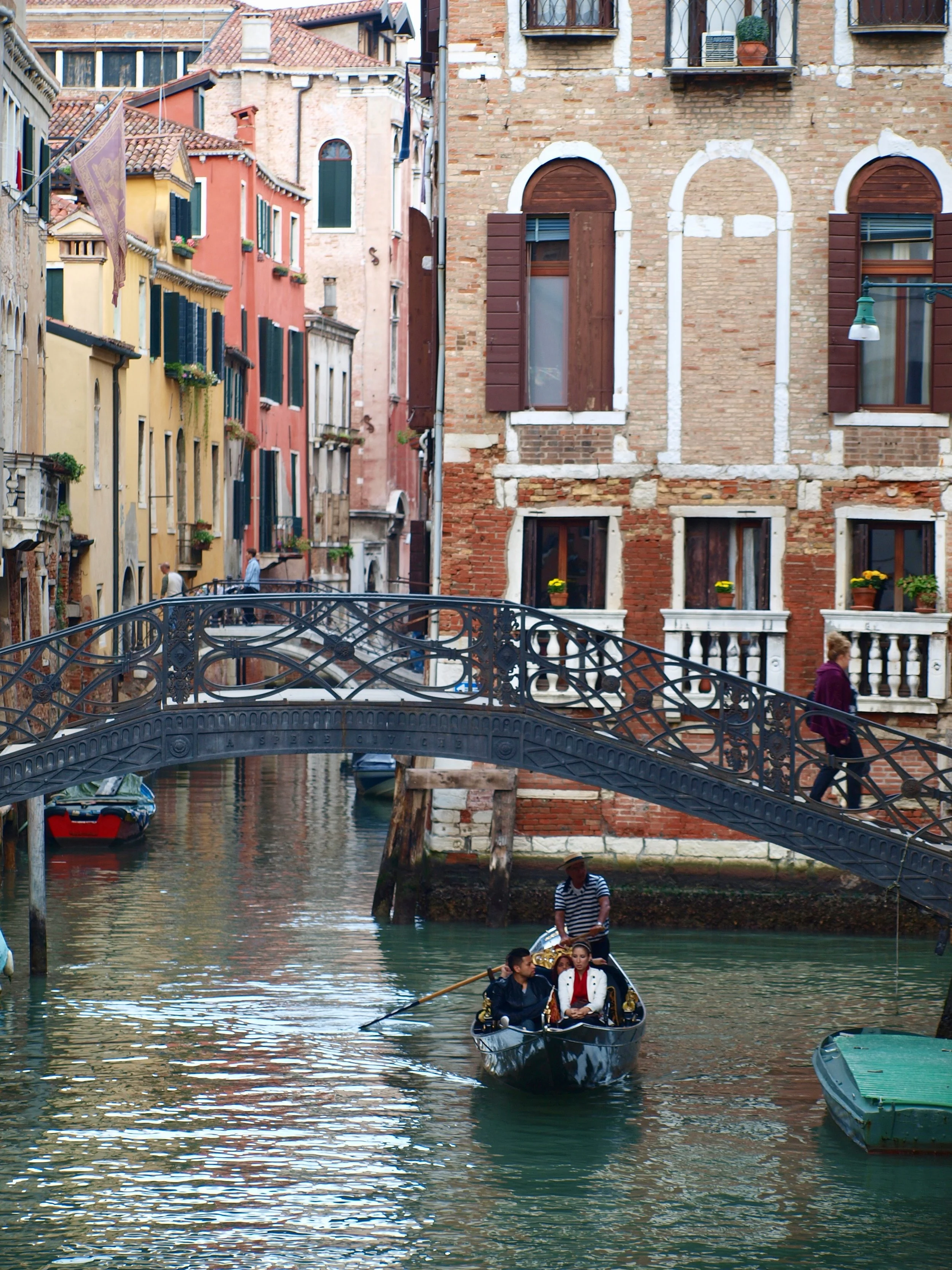 Une scène de Venise avec des bâtiments colorés en briques et un canal. Une gondole avec deux personnes, un homme et une femme, navigue sur l'eau, et un pont en fer ornée traverse le canal. Deux personnes se tiennent au bord de l'eau, près d'un bâtime