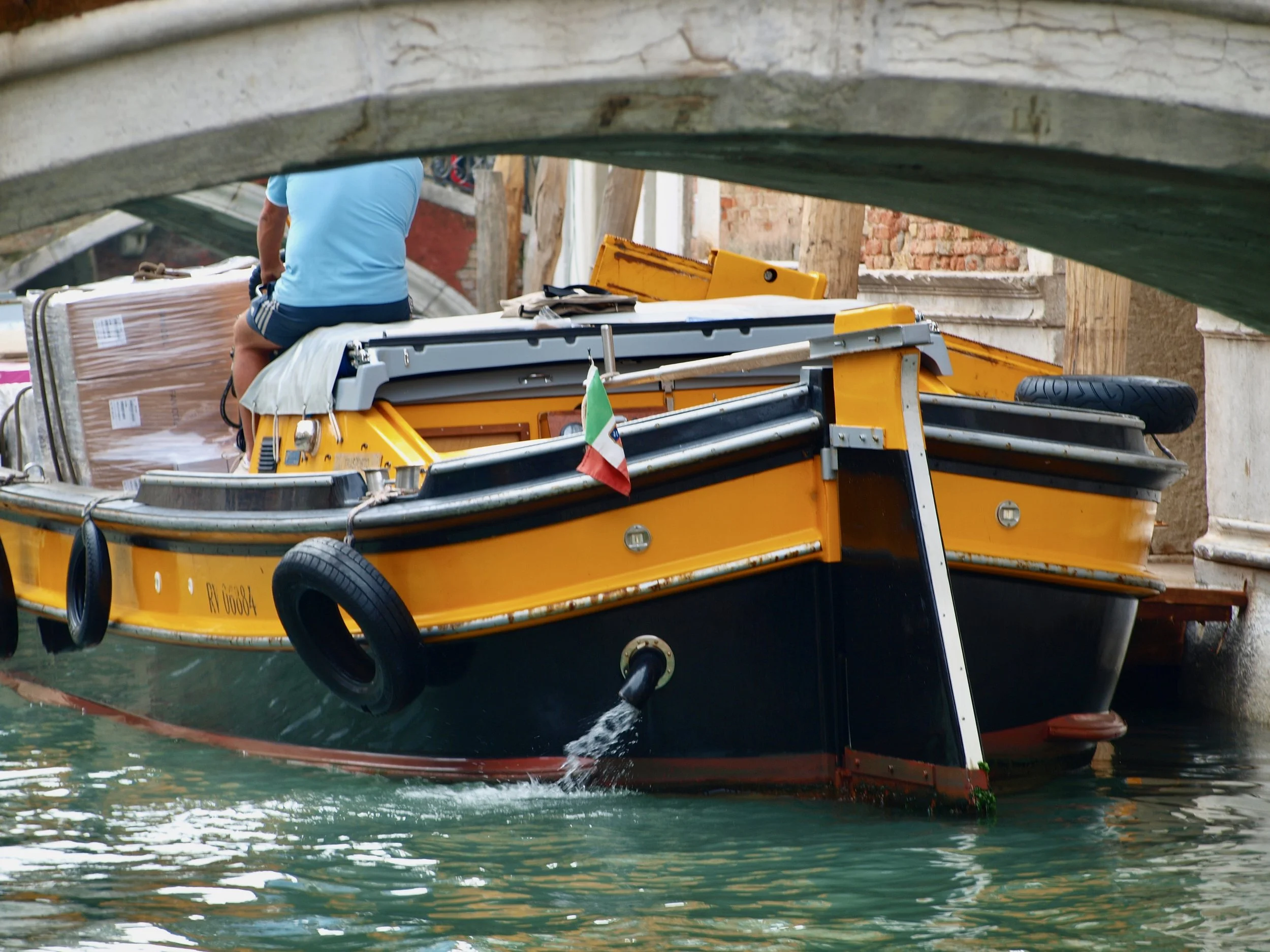 Un bateau jaune naviguant dans un canal, avec une personne à bord, vue partiellement. La photo est prise sous un pont en pierre.
