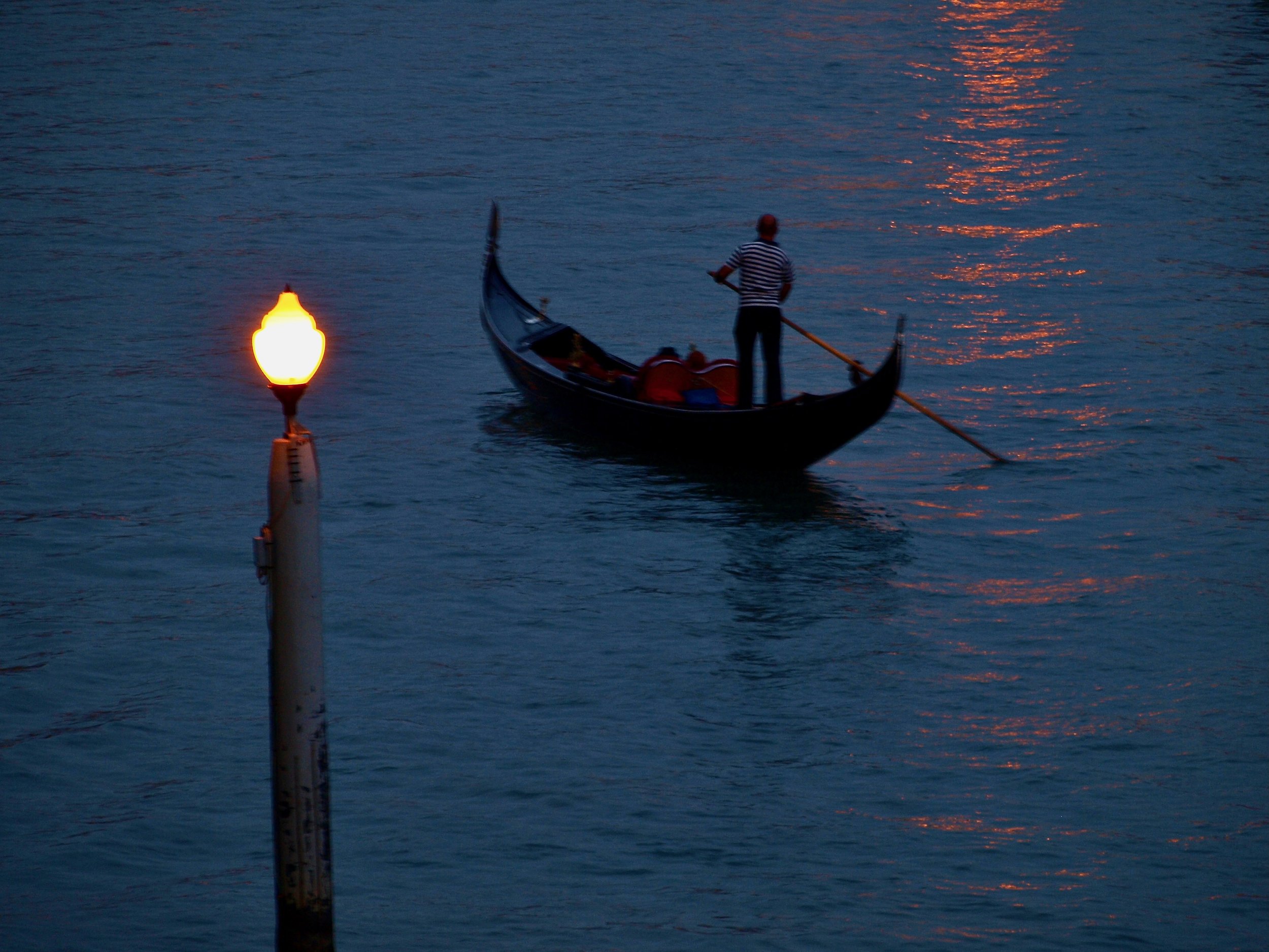 Une personne en gondole sur l'eau au crépuscule, avec un phare ou une balise lumineuse à gauche.