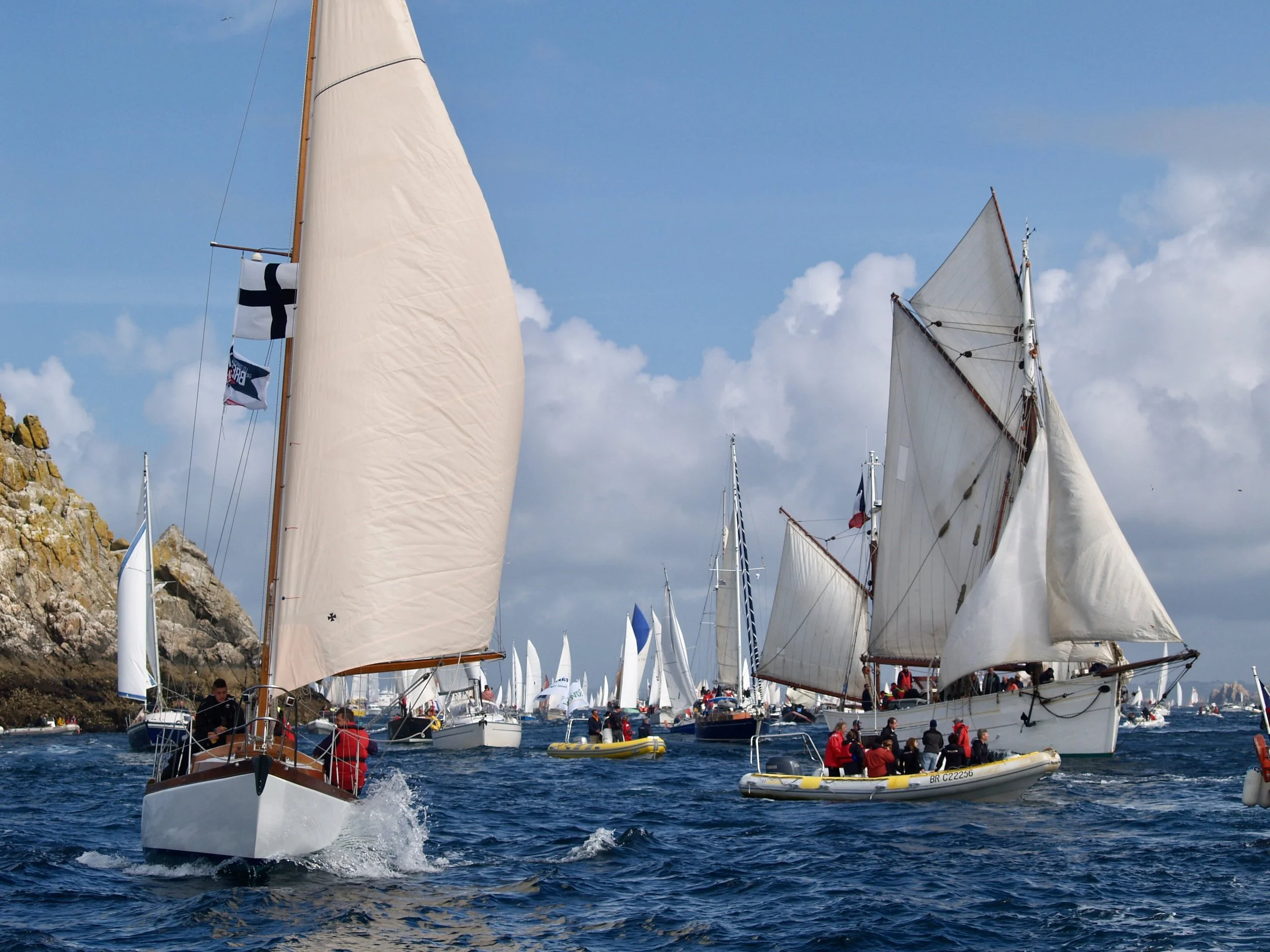 Plusieurs voiliers avec des voiles blanches naviguent dans une mer agitée, certains avec des foules de personnes à bord.