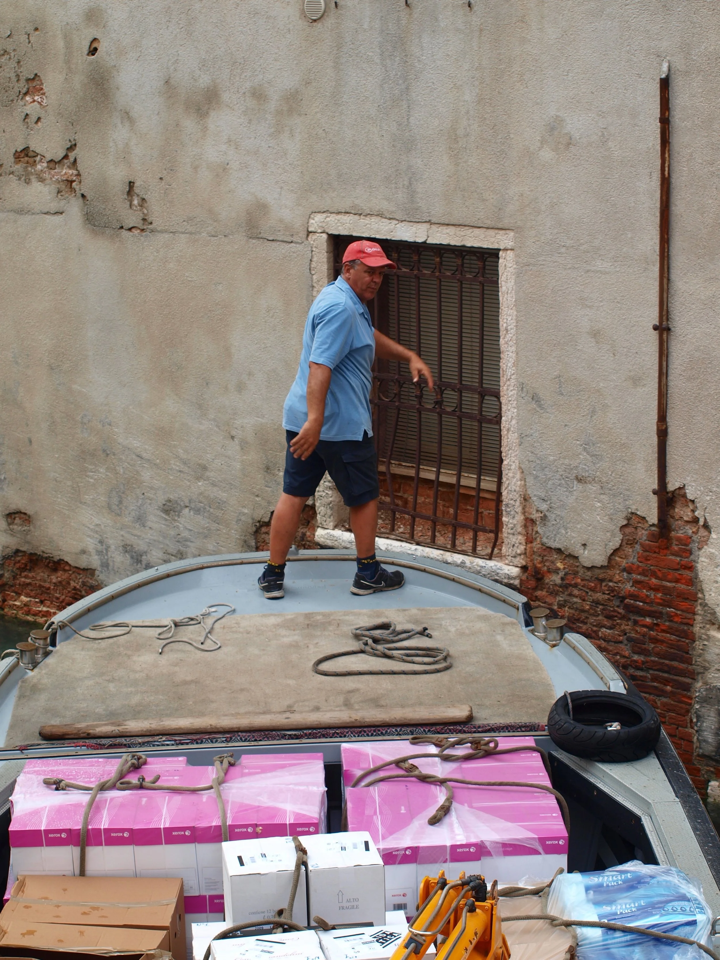 Un homme debout à côté d'une grande boîte recouverte de cordes, avec des appareils photo et des cartons autour, contre un mur en briques et ciment.