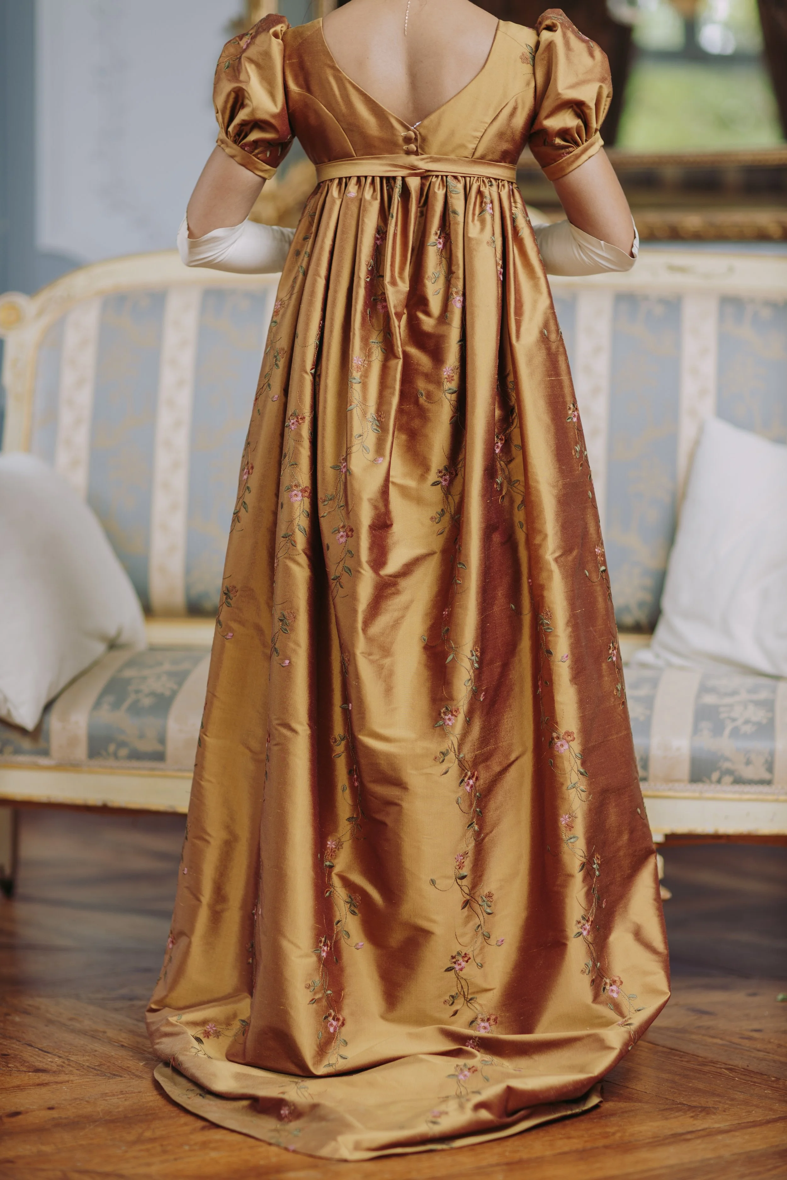 Une robe de soirée en satin doré avec des broderies de fleurs, portée par une personne avec des bras recouverts de gants blancs, dans un intérieur décoré avec un canapé et un tableau en arrière-plan.