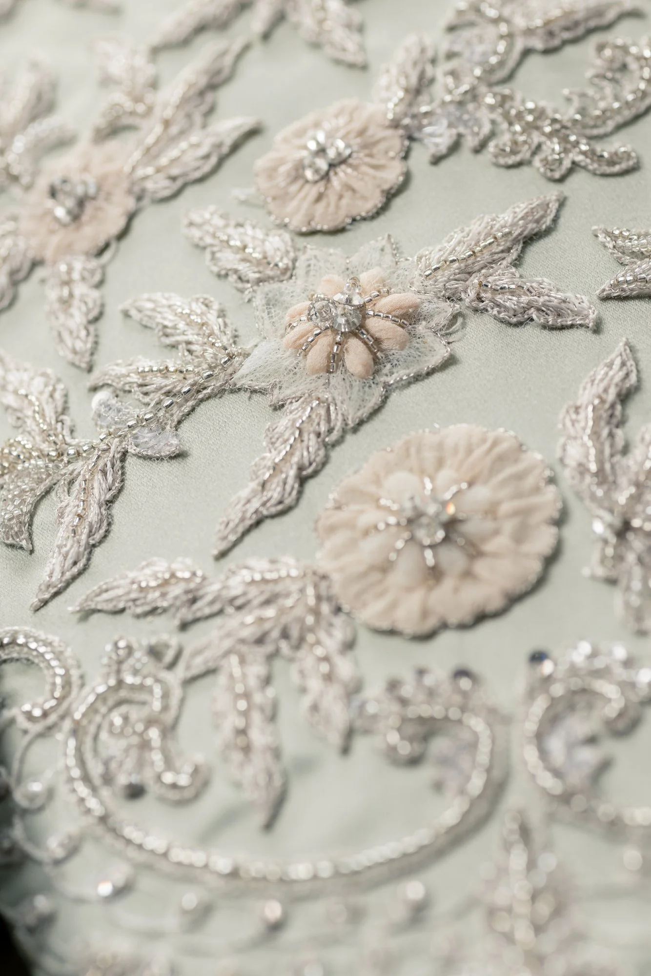 Broderie délicate avec motifs floraux et feuilles, ornée de perles et de sequins dans des tons dorés et ivoire sur un tissu pastel. 