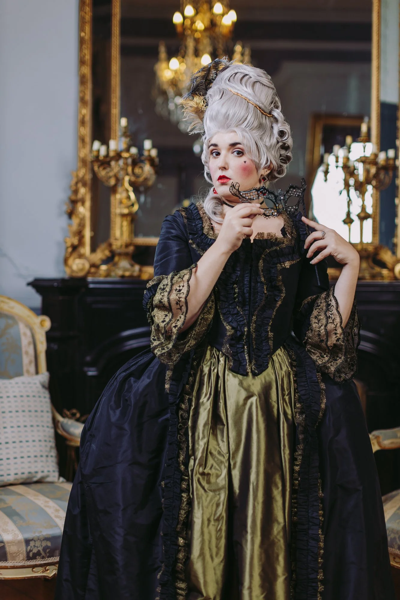 Une femme habillée en robe à la française historique rococo 18ème siècle historique, avec une perruque poudrée, tenant un masque en forme de papillon dans la main, dans une pièce élégante avec un grand miroir doré et un chandelier