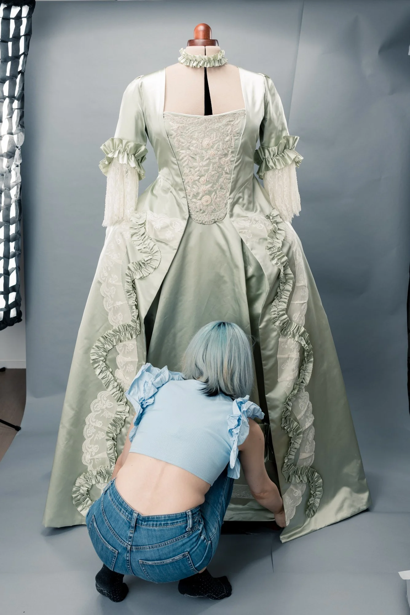 Une couturière assise, arrange une robe historique ancienne robe à la française en satin de soie vert avec des détails en dentelle rococo, probablement en restauration ou en préparation pour une exposition ou un shooting photo.