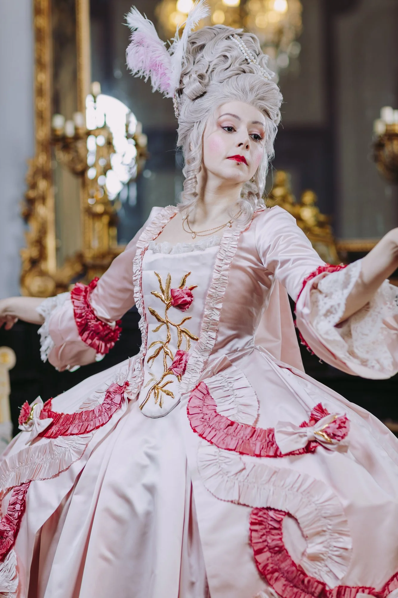 Une femme habillée en robe à la française historique du XVIIIe siècle rococo, ornée de broderies détails couture et de rubans, portant une coiffe élaborée avec des plumes blanches et des accessoires.