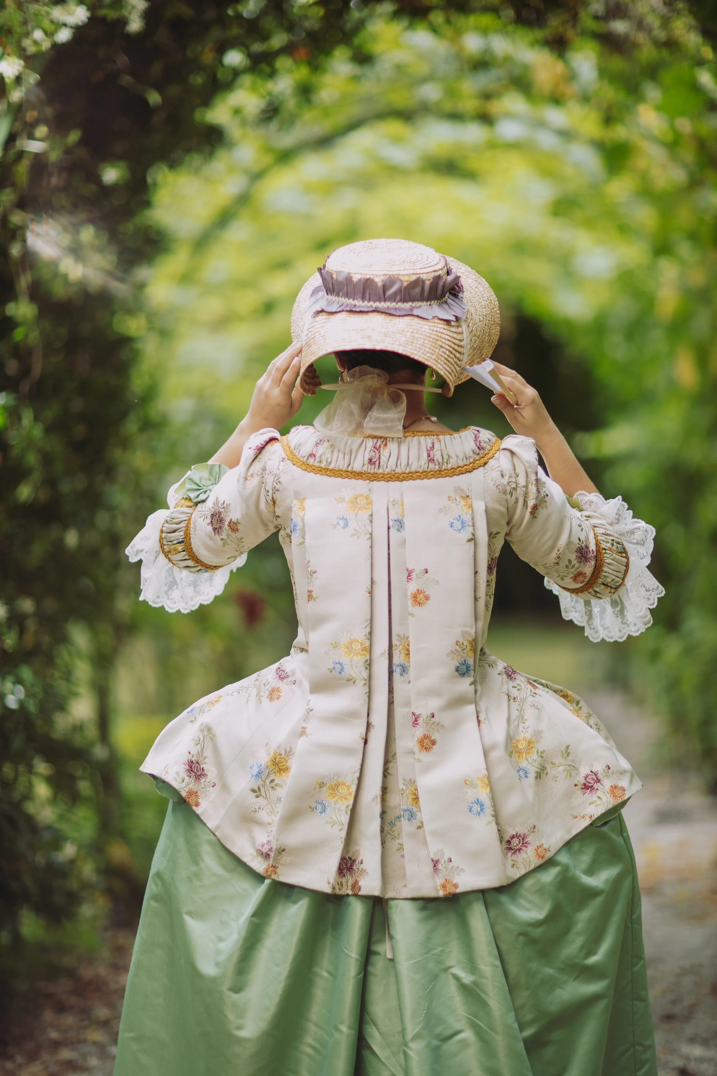 Une personne portant une robe de style historique pet en l'air plis watteaux 18ème siècle rococo avec des engageantes en dentelle, un chapeau en paille décoré d'une bande en tissu violet, dans un cadre naturel avec des arbres verts en arrière-plan.