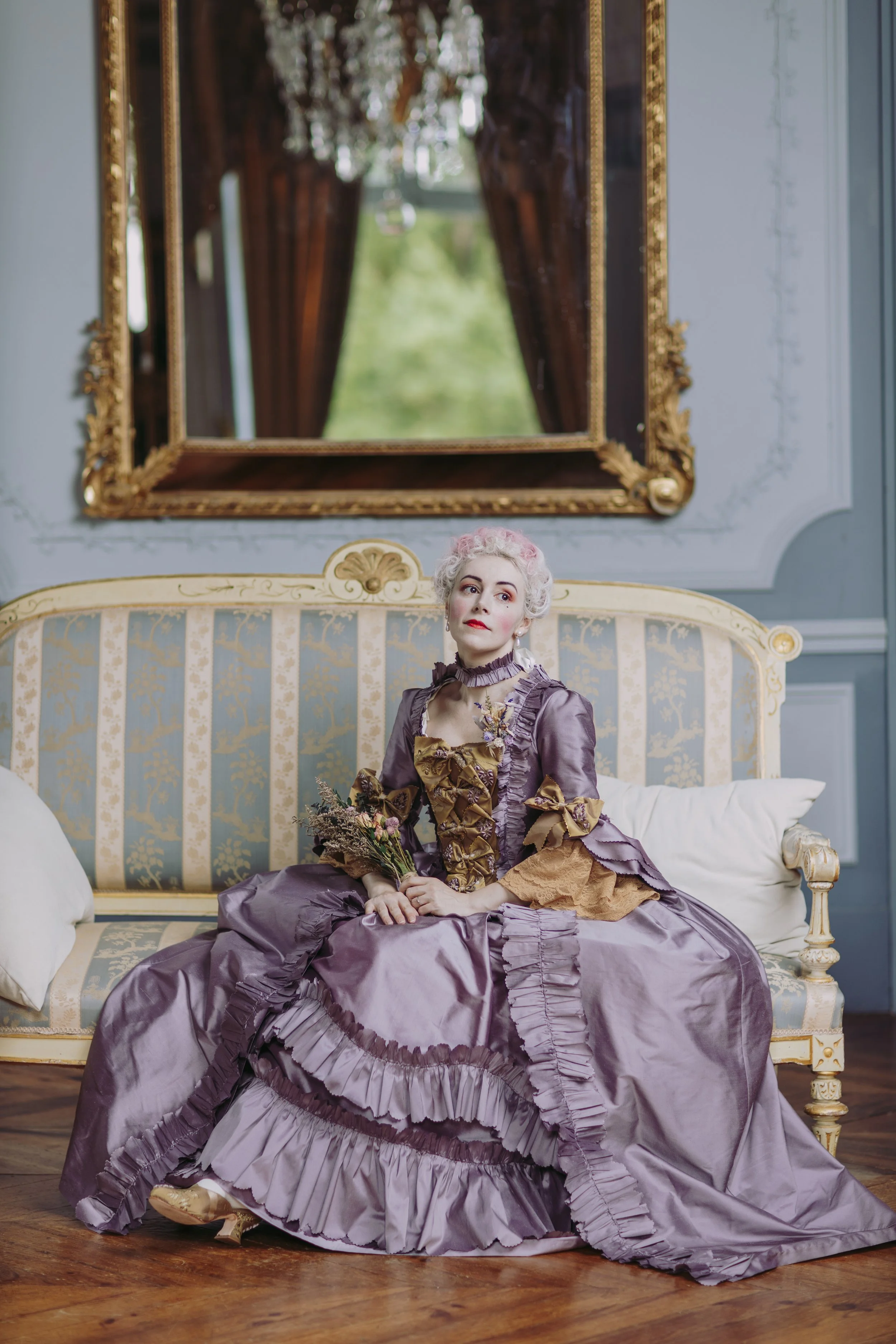 Une femme dans une robe historique à la française de style rococo 18ème siècle assise sur un canapé doré. Elle tient un bouquet de fleurs et pose dans une salle ornée d’un cadre doré et d’un grand miroir.