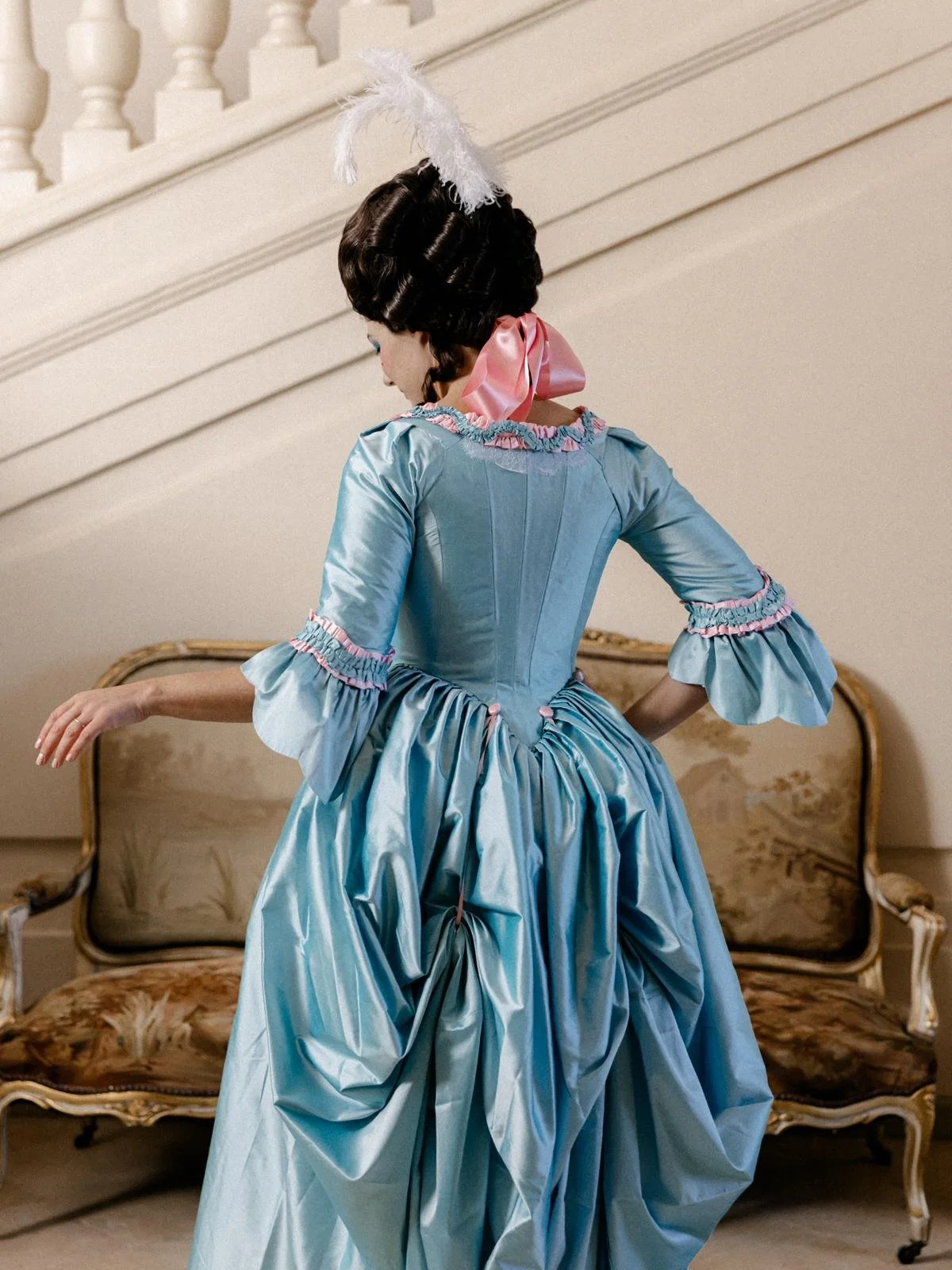 Femme avec une robe à l'anglaise historique en soie bleu 18ème siècle détails couture rococo, un nœud rose dans les cheveux et un éventail, vue de dos, dans un intérieur élégant.