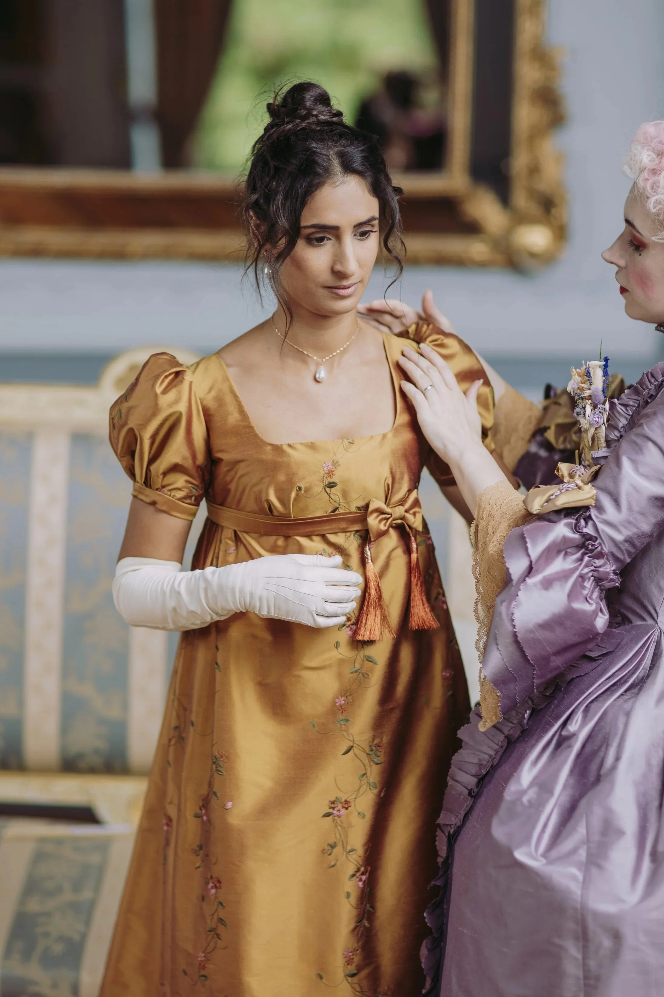 Deux femmes habillées en style premier empire bridgerton, une en doré et l'autre en violet, en train de se faire aider à enfiler une chemise ou un vêtement. La scène se passe dans une pièce élégante, avec un miroir doré en arrière-plan.