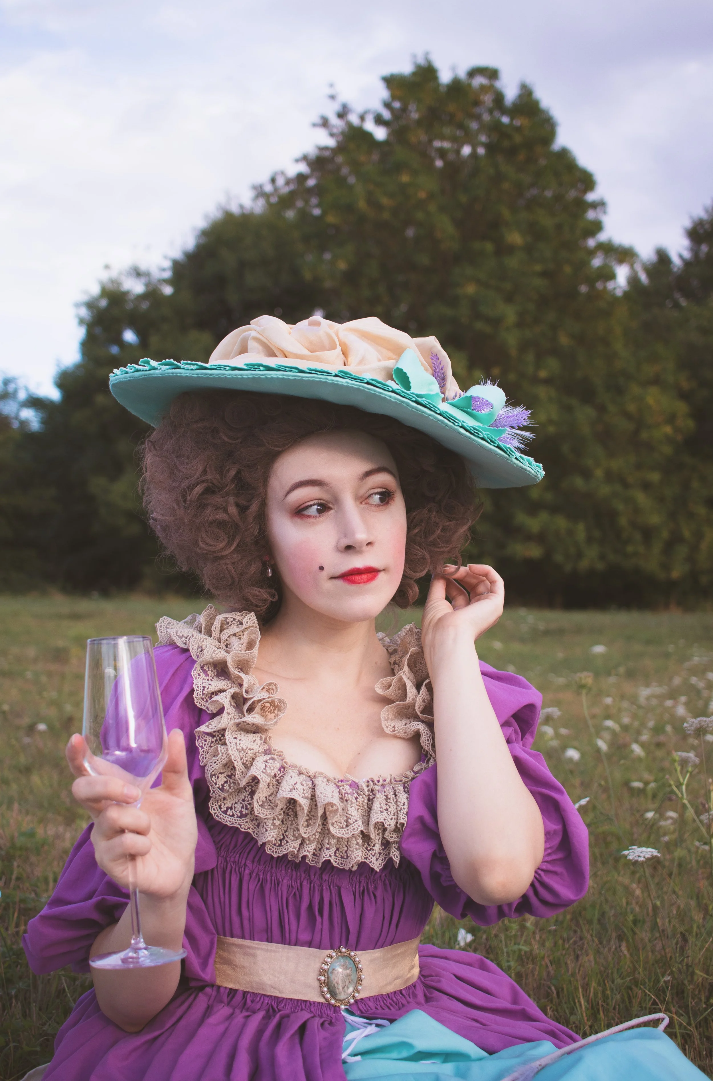 Une femme portant un grand chapeau en décoration florale, une chemise à la reine cousue mains 18ème siècle historique violet avec une ceinture en perles, tenant un verre à vin violet dans un champ avec des arbres en arrière-plan.