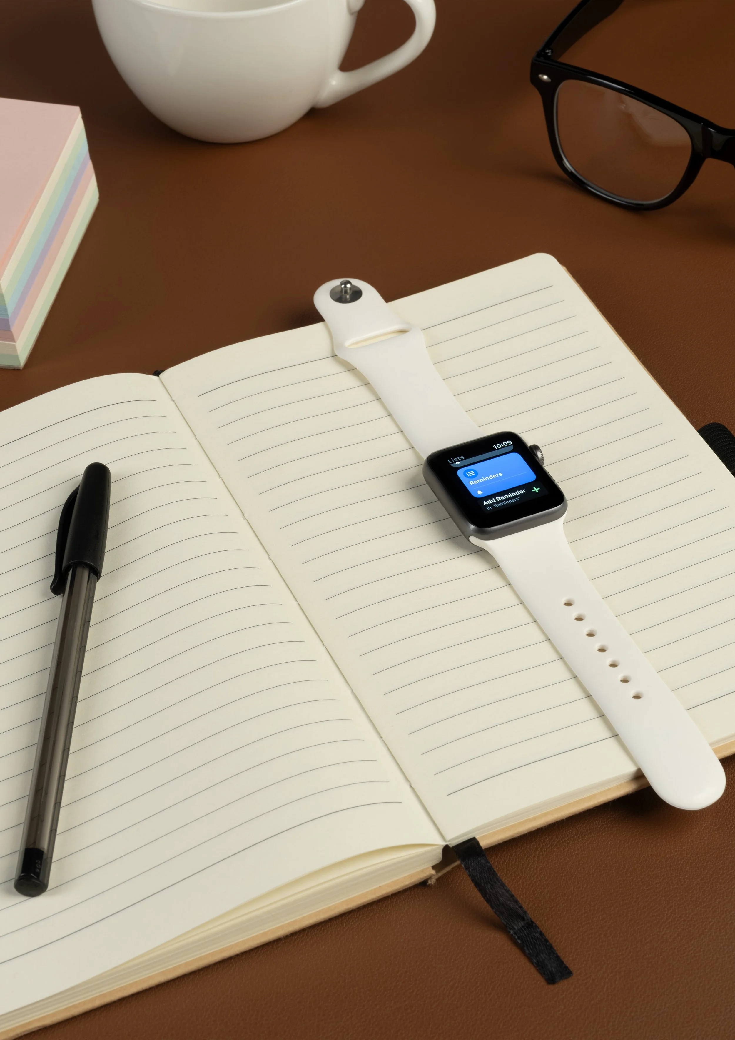A brown desk with a white ceramic mug, a stack of pastel-colored sticky notes, a black pen, an open notebook with lined pages, a smartwatch displaying reminders, and a pair of black glasses.