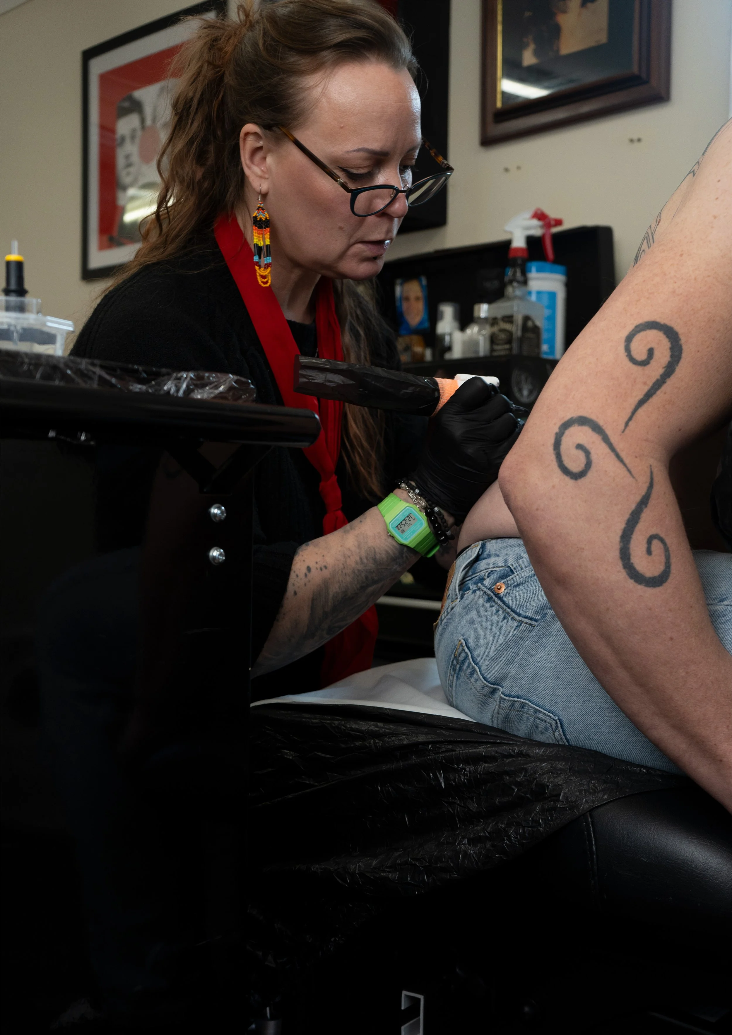 A woman with long brown hair, wearing glasses, beaded earrings, a red lanyard, and black gloves, tattooing a person's arm with black ink in a tattoo studio. The person getting tattooed has a large tattoo on their arm and is wearing ripped jeans.