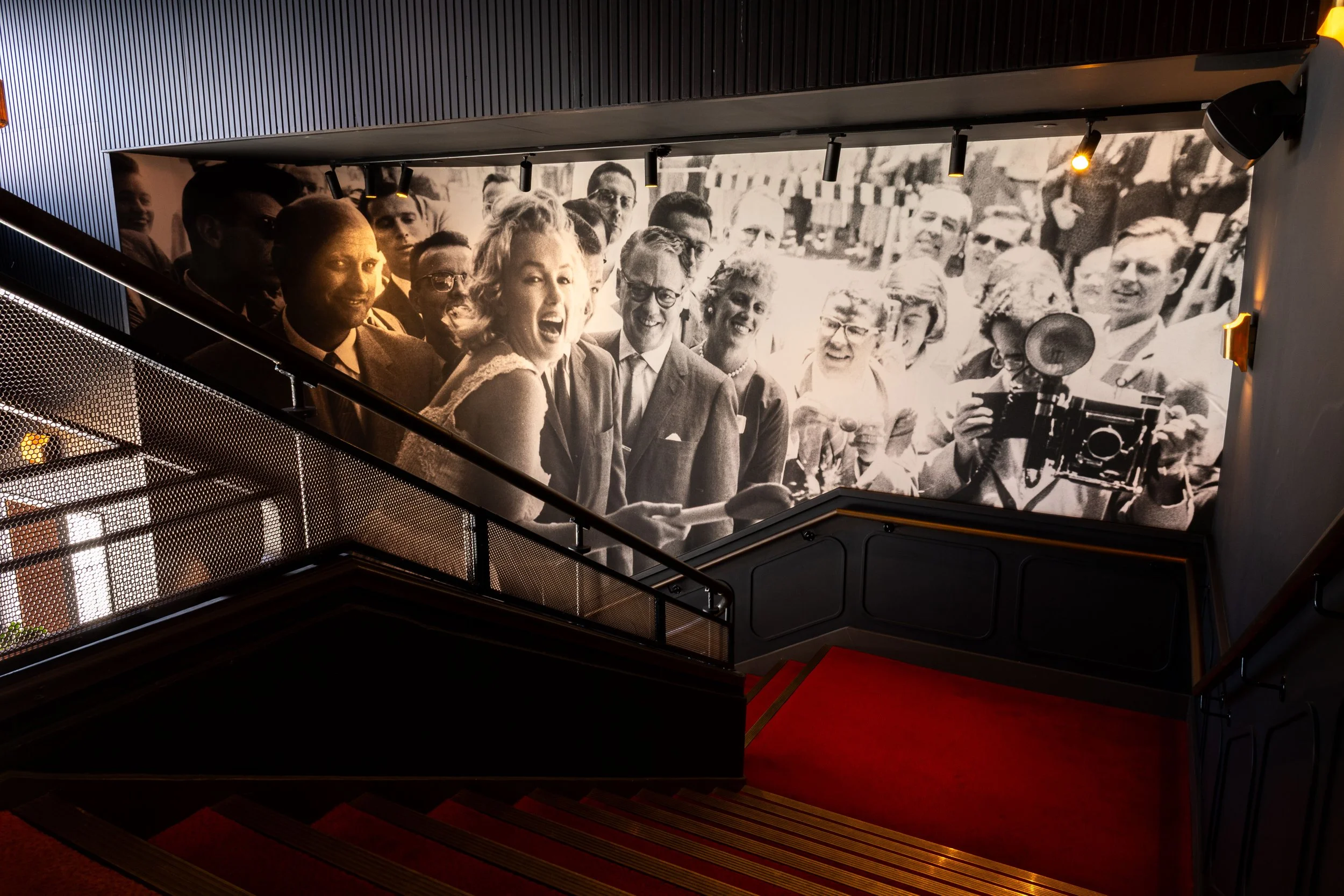 A large black-and-white photograph of a group of smiling people, including men and women, some with glasses, in a public outdoor setting, displayed on a wall inside a building.