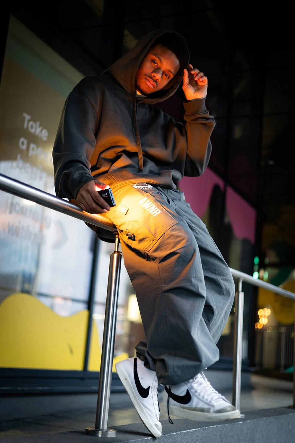 A young man in a black hoodie and gray cargo pants with white Nike sneakers poses on a railing at night.