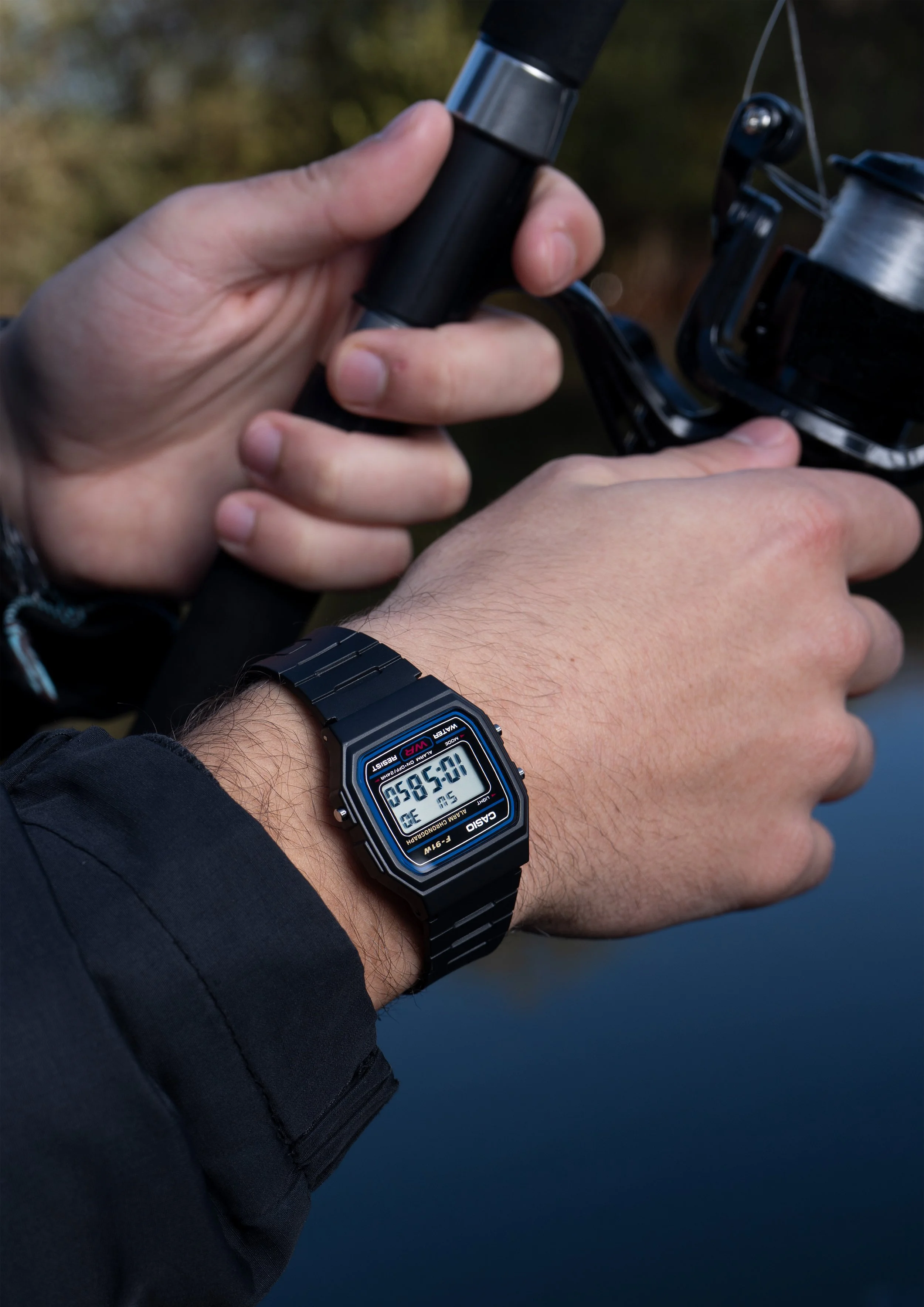 Close-up of a person's hand holding a fishing rod and reel, with a digital Casio watch showing 9:55:50 on their wrist, against a blurred outdoor background.