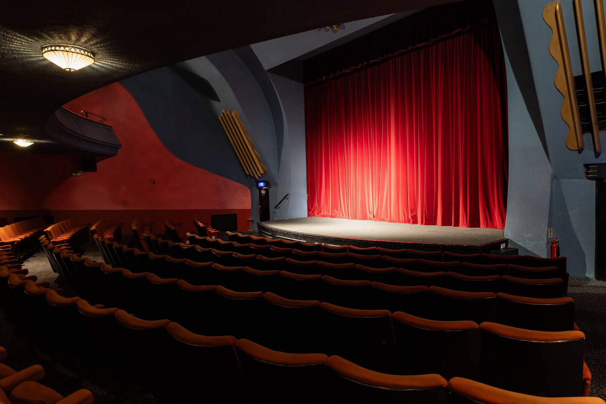 Empty theater with a stage covered by a red curtain and surrounding seats.