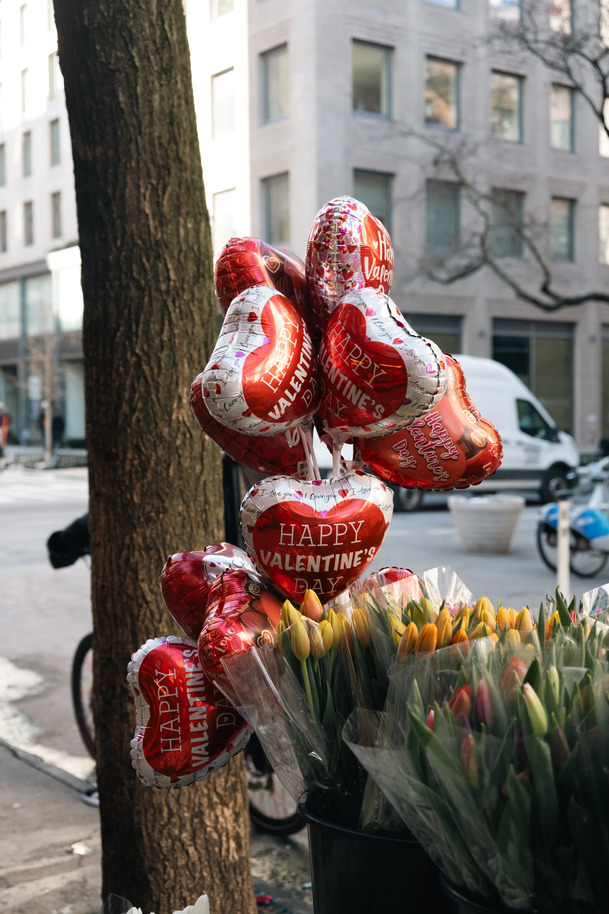 A bunch of Valentine's Day themed balloons with the message 'Happy Valentine's Day' in red and white, displayed next to a bouquet of yellow tulips wrapped in plastic, outdoors on a city street.