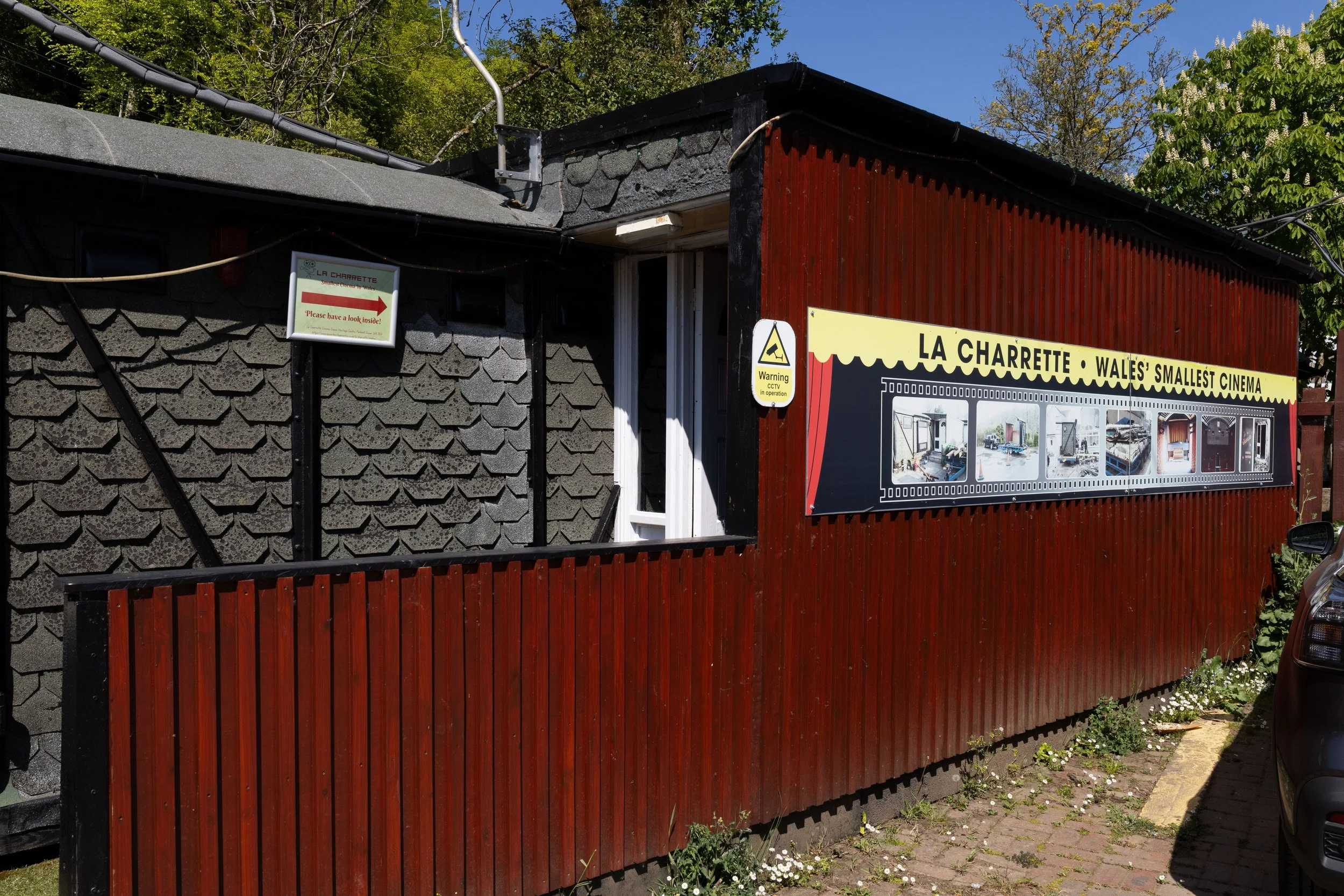 An outdoor scene featuring a small building with a blend of stone and wooden exterior. The building has various signs, including one indicating it is Wales' smallest cinema called La Charrette. There is a red wooden fence in front of the building, gr