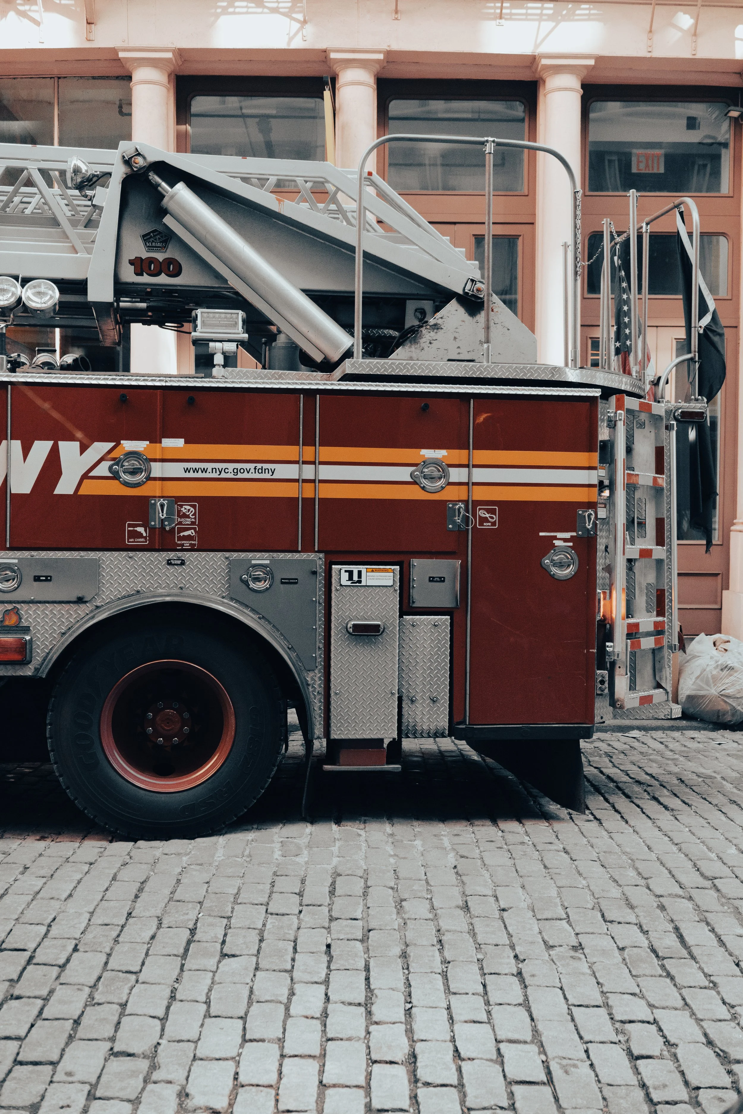 The image shows a close-up side view of a red fire truck with silver and black equipment, parked on a cobblestone street outside a building with large glass windows and white pillars.