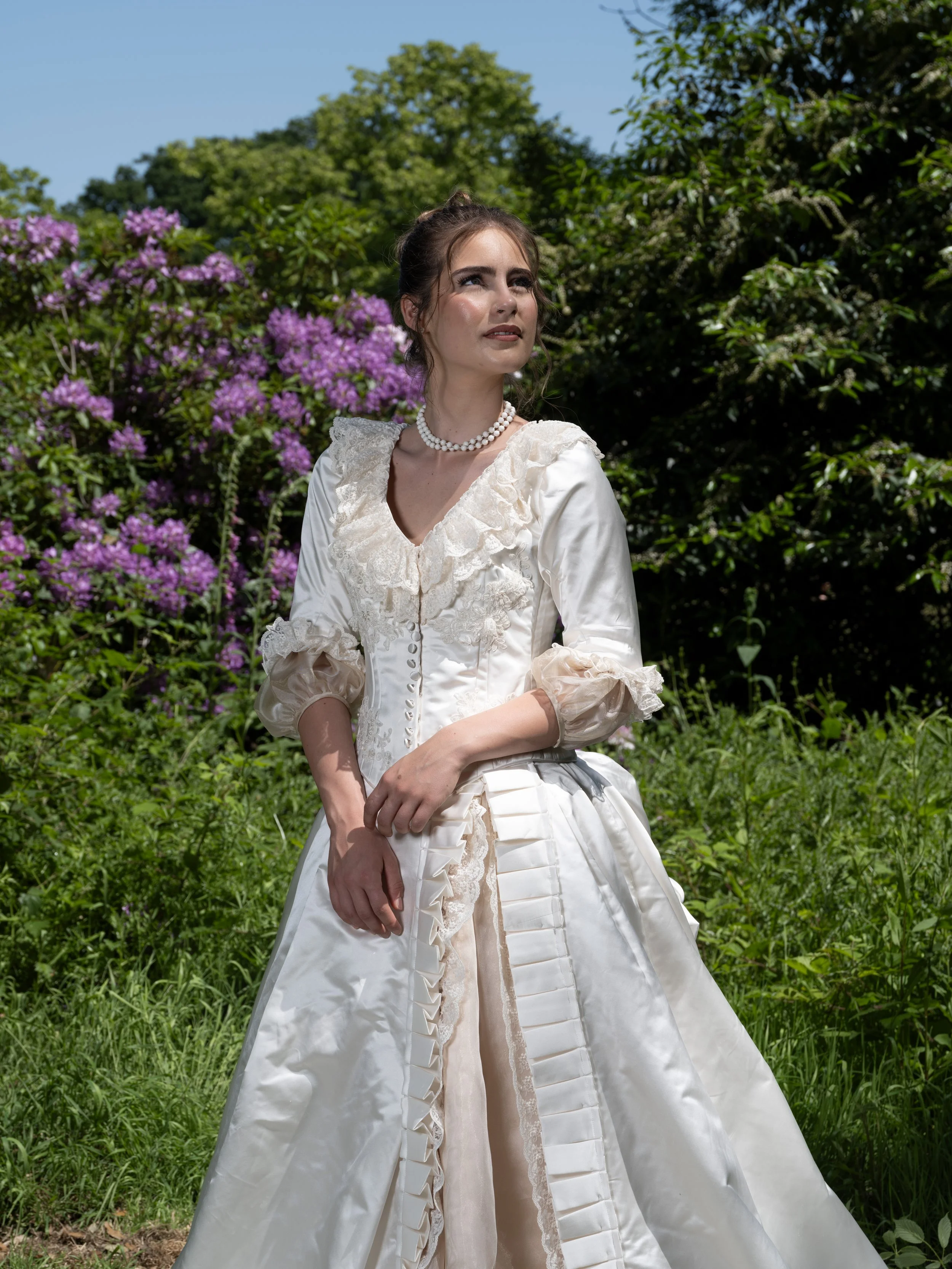 A woman in a vintage white dress with lace details and pearl necklace standing outdoors among green shrubbery and purple flowering plants under a clear blue sky.