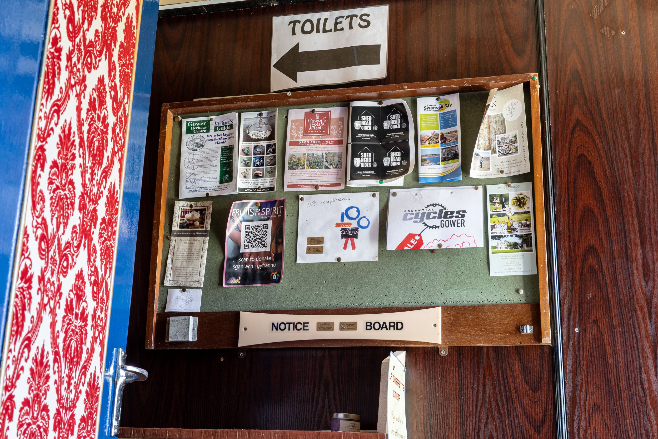 A wooden bulletin board displaying various flyers and notices, with a large white sign above it pointing left that says 'TOILETS'.