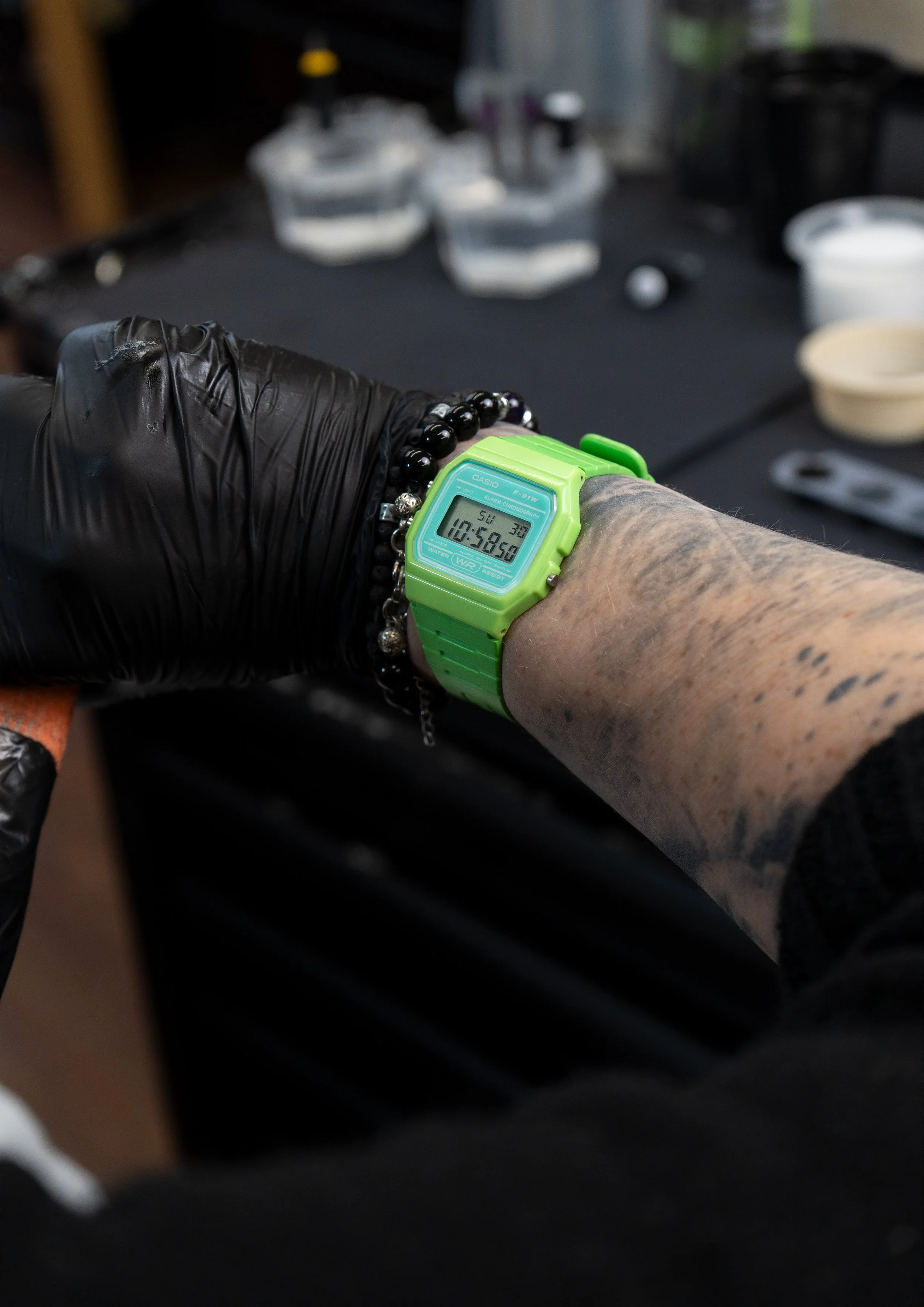 A person's arm wearing a green Casio digital watch and a black beaded bracelet, with the background showing a blurred table with containers and tools.