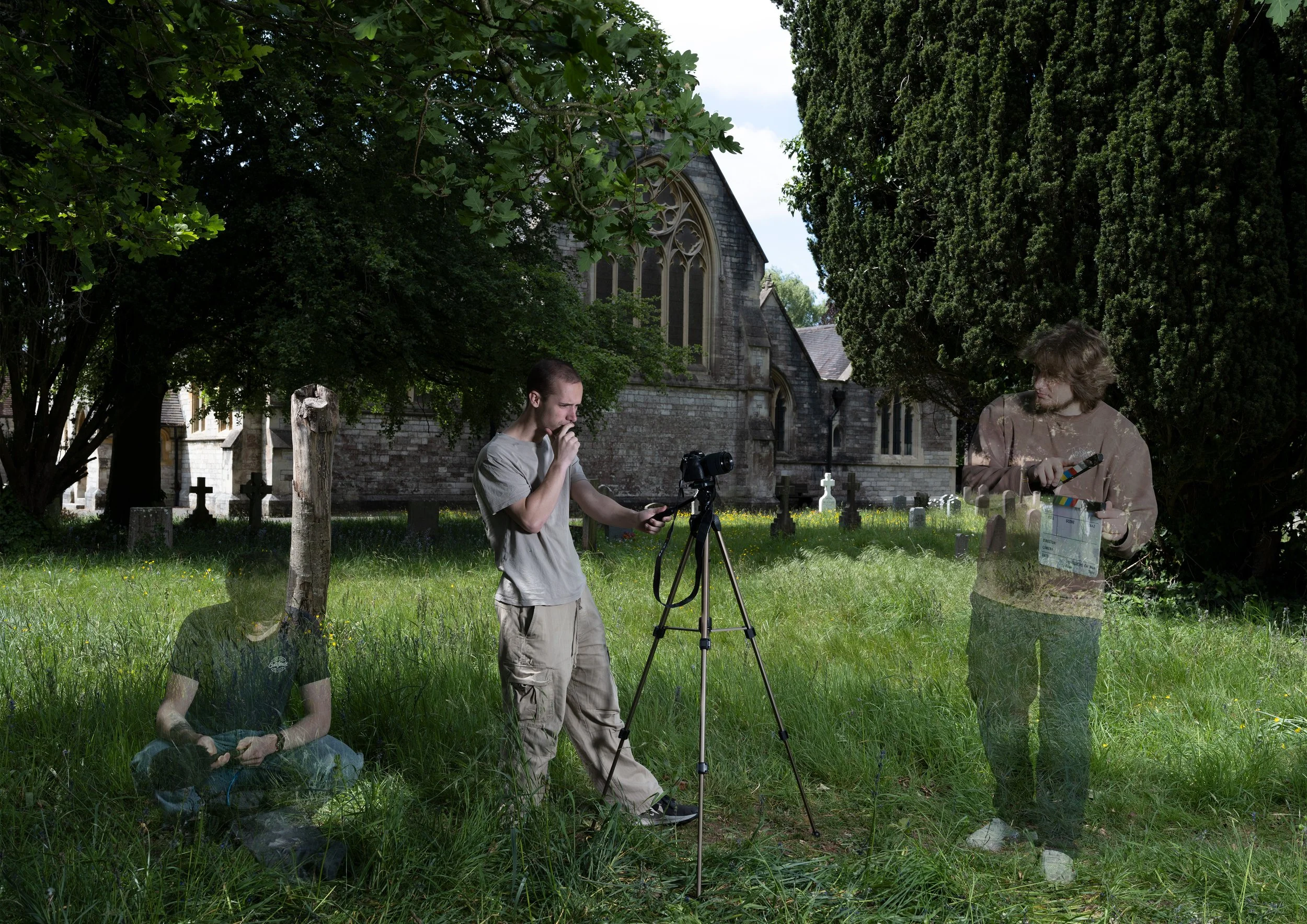 Two young men are in a cemetery with old gravestones and large trees. One is sitting on the ground, and the other stands behind a camera on a tripod, looking at his phone. The scene is partly overlapped by a transparent layer showing the same scene i