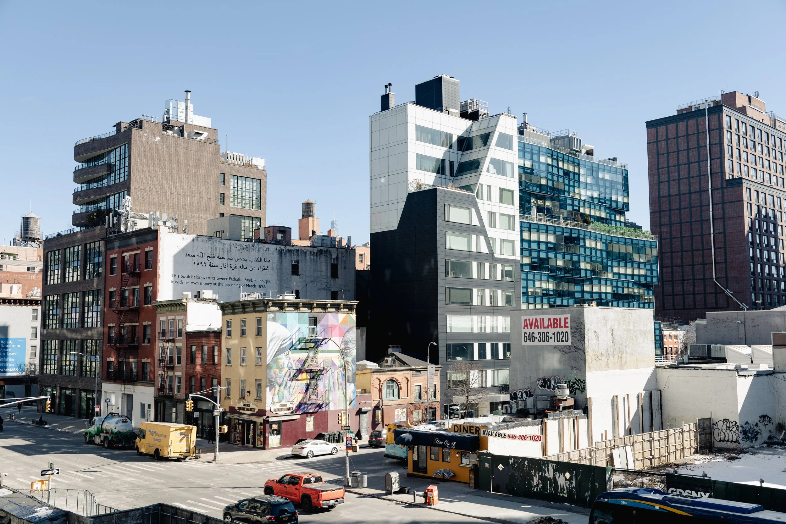 A cityscape with tall modern buildings in the background and smaller older buildings in the foreground. The scene includes parked cars, a street intersection, and some graffitied walls. Clear blue sky overhead.