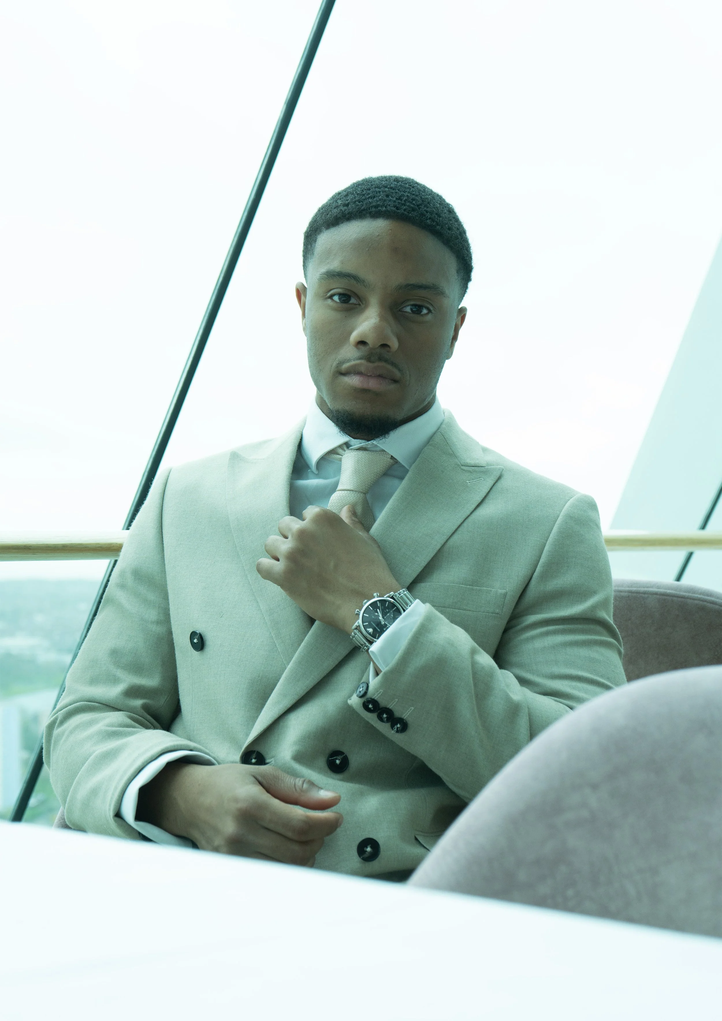 A young man in a beige suit and tie adjusting his collar, wearing a watch, standing indoors near a large window with a modern architectural exterior visible.