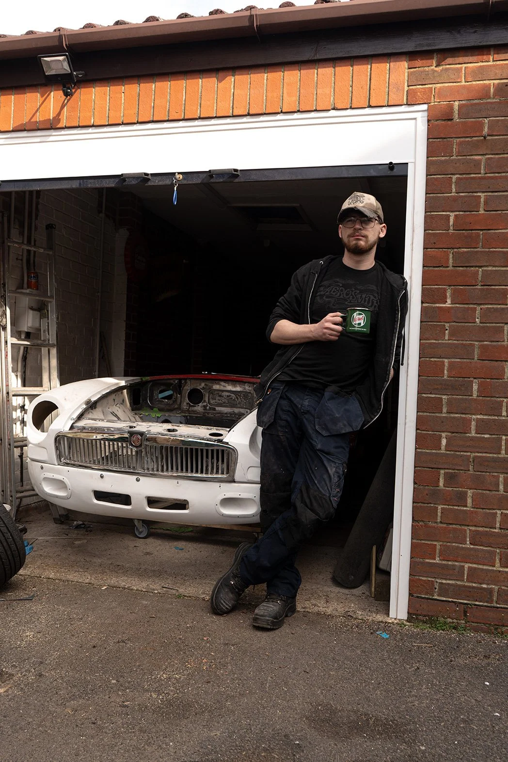A man in casual clothes, including a cap and glasses, stands outside a garage in front of an unfinished vintage car body. He is holding a green mug and leaning against the garage's brick wall. The garage has a brick exterior with a brown tiled roof a
