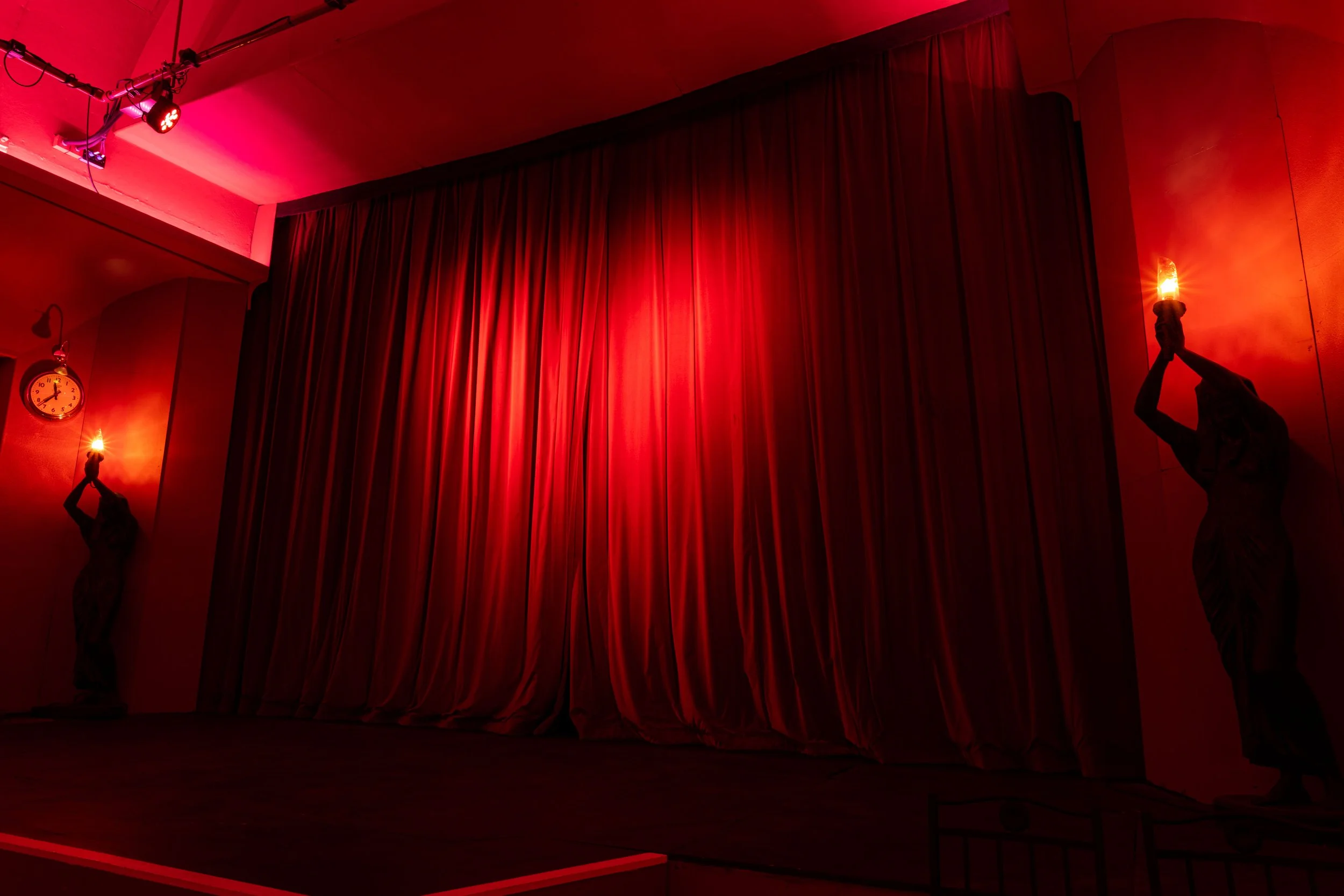 The image shows a stage with closed red curtains, dim red lighting, a clock on the wall, and a statue of a person holding a torch, all illuminated by warm, low-intensity lights.