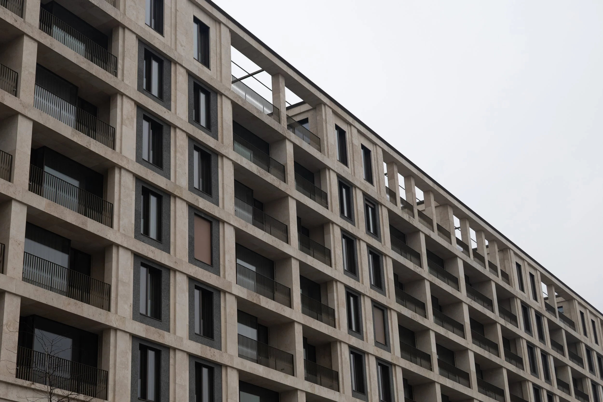 A multi-story modern residential building with repetitive window and balcony patterns, beige stone facade, and black balcony railings, under a gray, overcast sky.