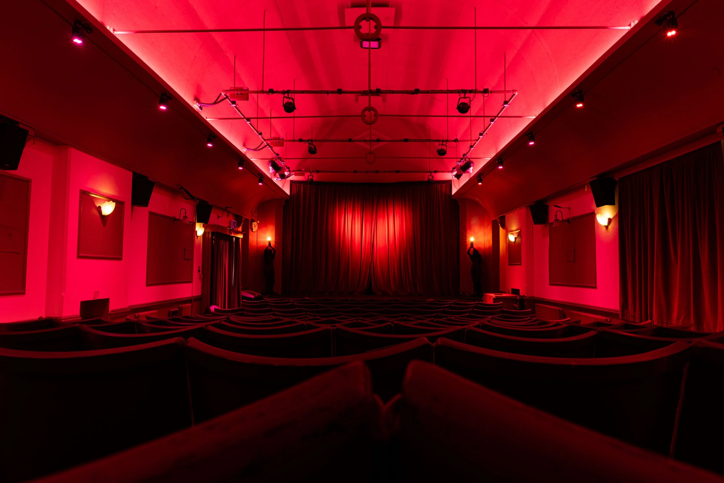 An empty theater with red lighting, red curtains at the stage, and rows of red seats.