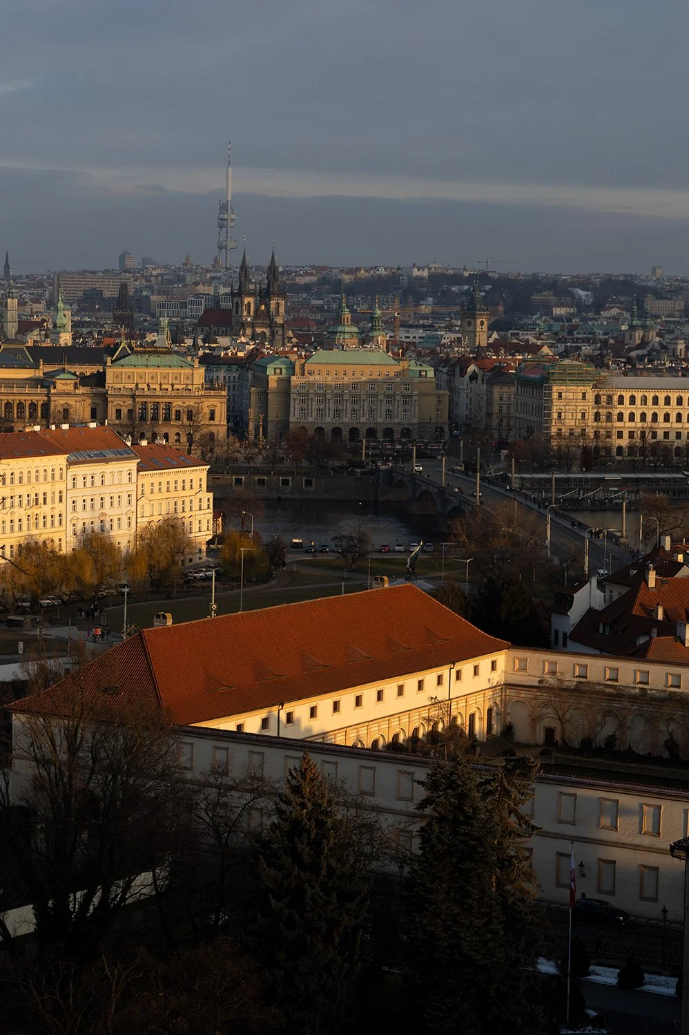 A cityscape of Prague, Czech Republic, featuring historic buildings, the Vltava River, bridges, and a television tower in the distance, during late afternoon or early evening.