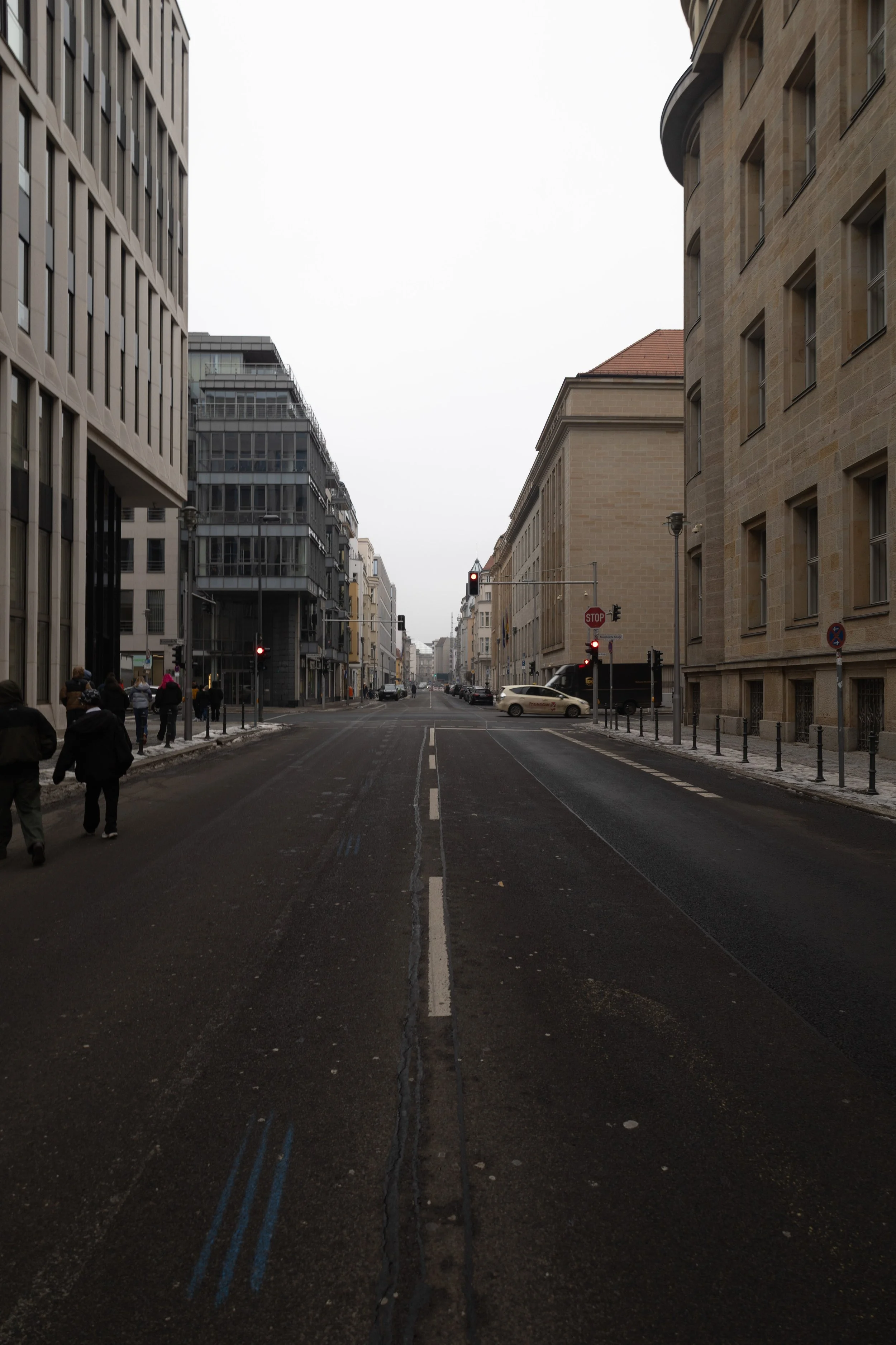 A city street view with modern and historic buildings on both sides, a traffic light showing red, a few cars, and pedestrians walking on the sidewalk on a cloudy day.