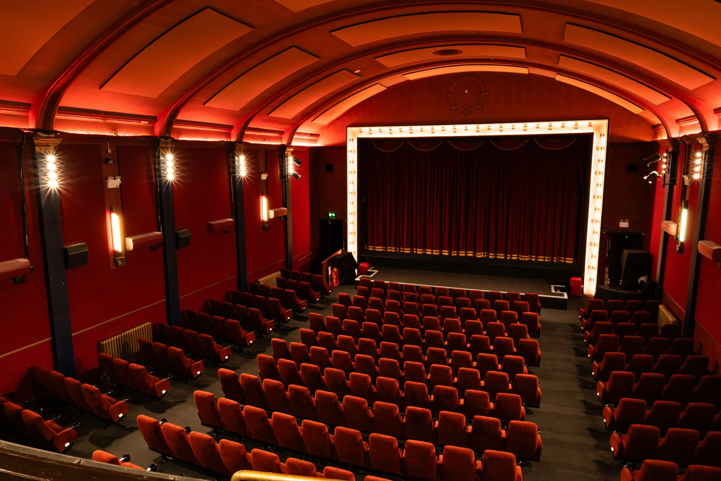 An empty theater with red velvet seats, a stage with a closed curtain, and warm lighting. The ceiling has a decorated pattern with glowing lights.