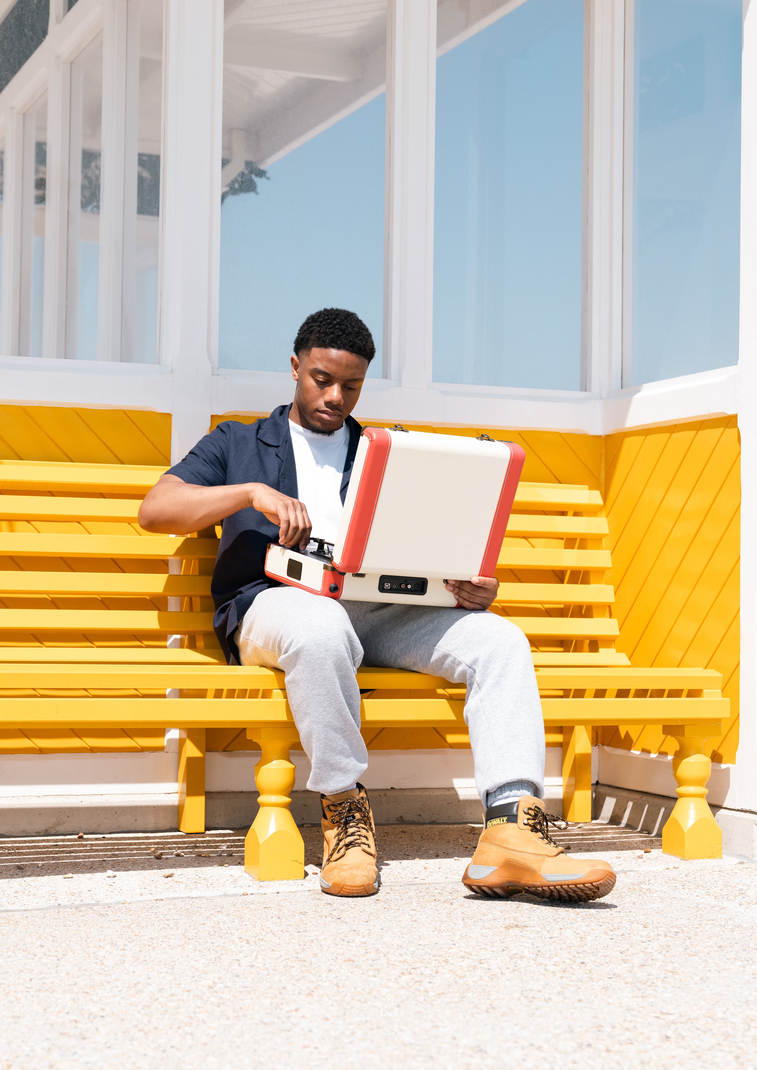 Young man sitting on a bright yellow bench outdoors, looking into a toolkit or case, wearing tan work boots, gray sweatpants, a dark shirt, and a white t-shirt, with a building with glass windows behind him.