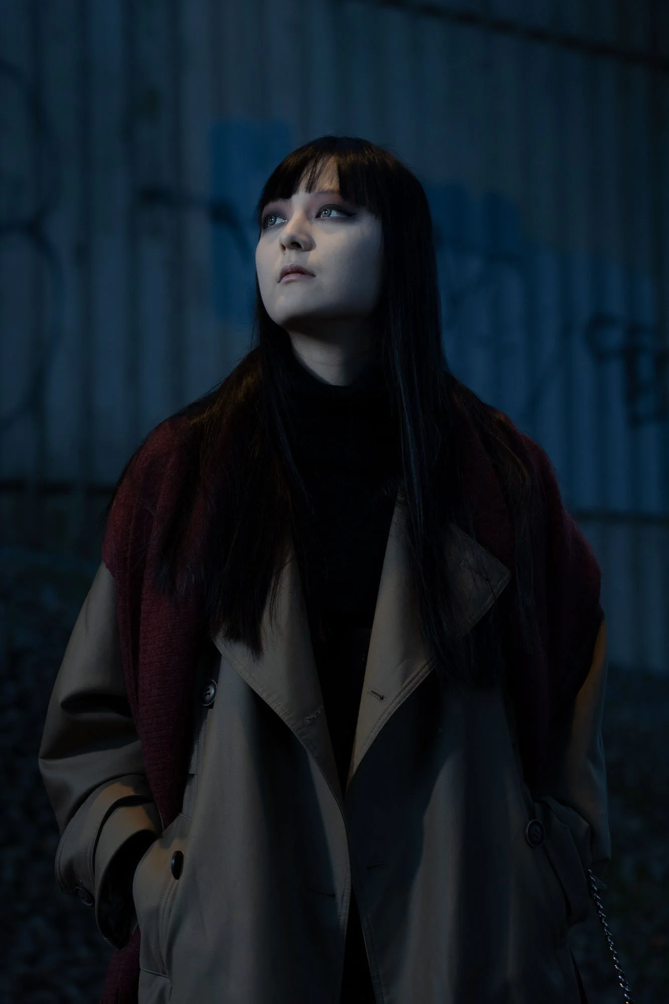 A woman with long dark hair and light skin looking to the side, dressed in a beige trench coat with a beige bag and a red garment over her shoulder, standing outdoors at night with a dark, blurred background.