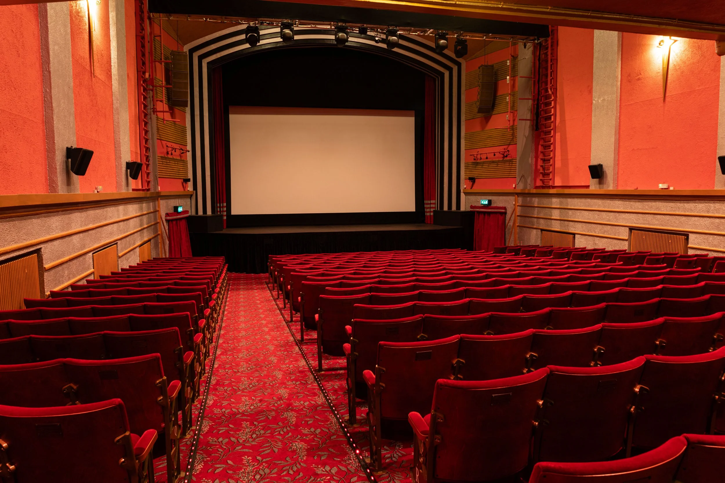 Inside a vintage theater with a large white movie screen, red velvet seats, and red walls. The theater has curtains on the sides of the screen and decorative lighting on the walls.